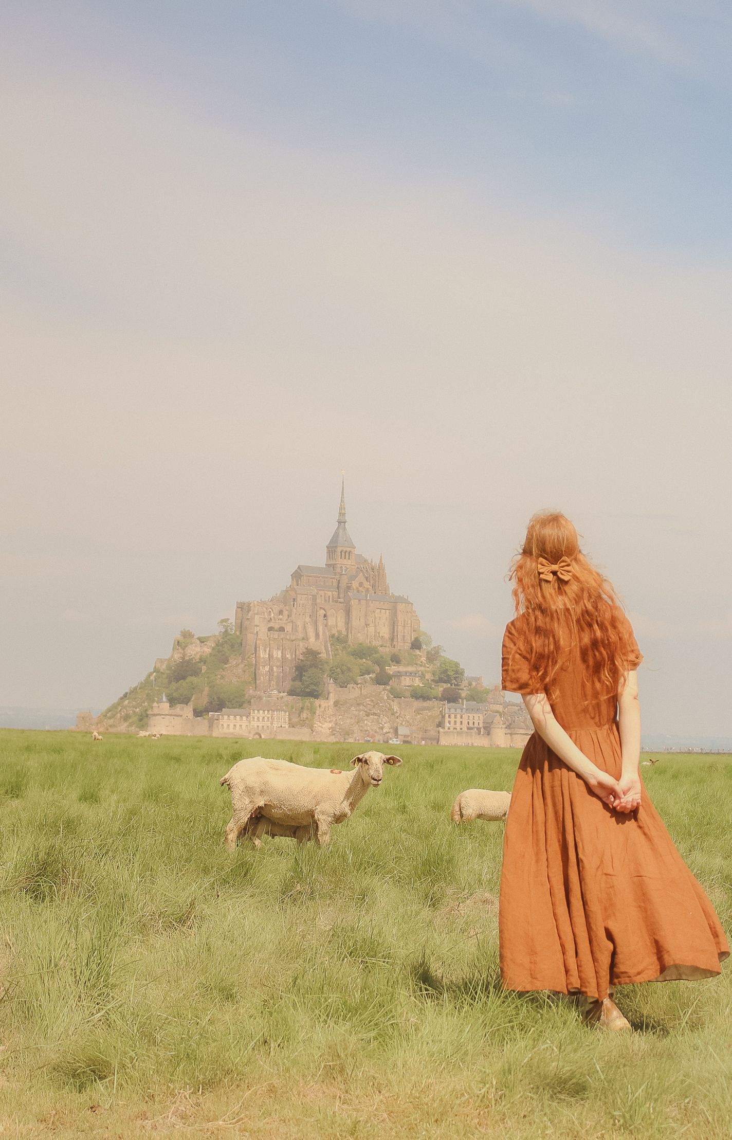 A woman with long ginger hair is standing in a field with sheeps and in the background you can see the Mont Saint Michel