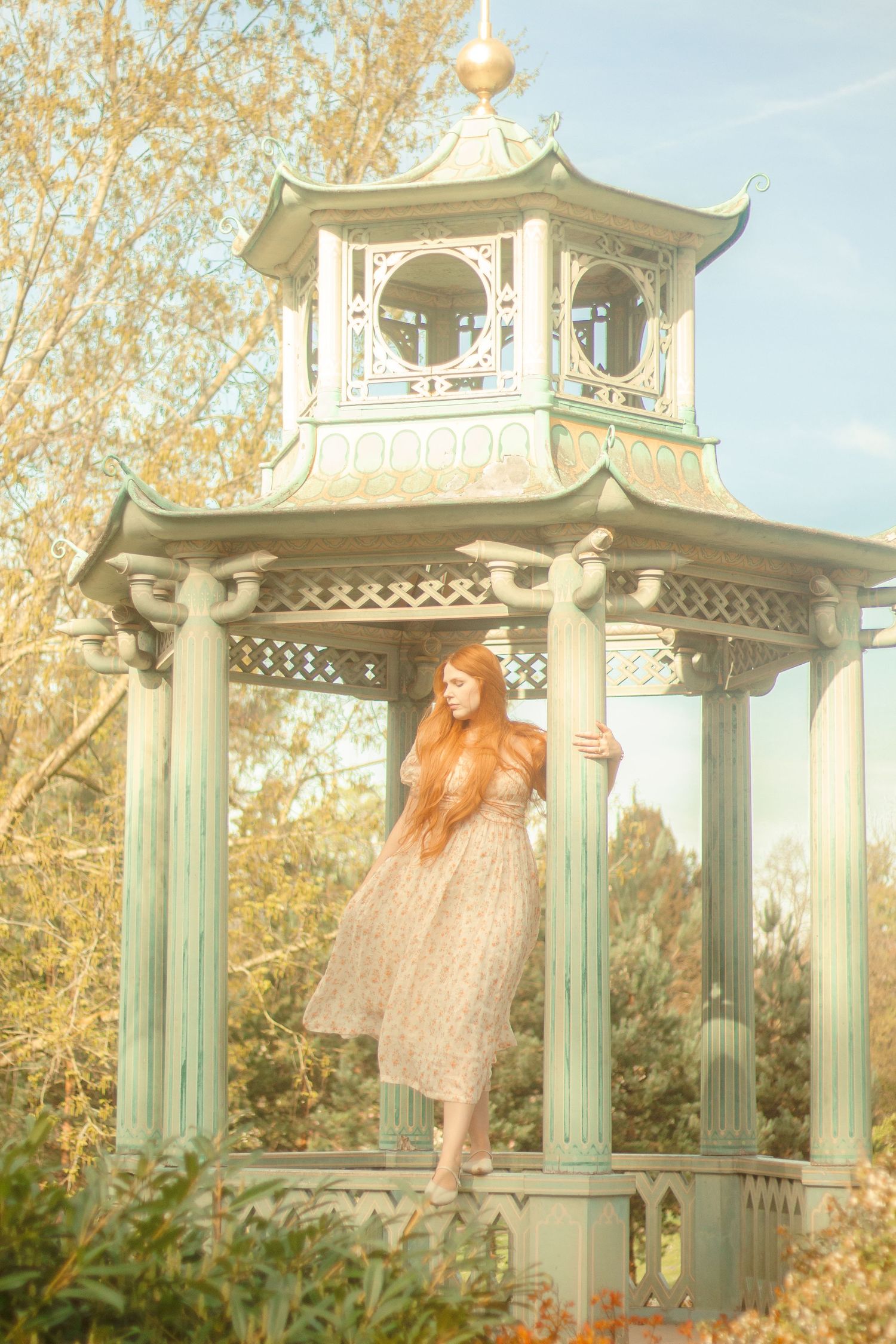 A woman with long red hair is standing in a decorative gazebo, wearing a floral dress. She is surrounded by a garden filled with flowers.