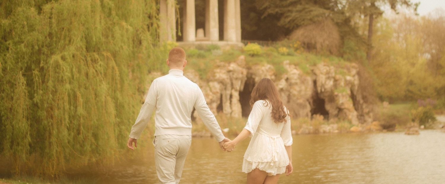 A couple is walking hand in hand, in the background there is a willow tree and a temple located in Bois de Vincennes.