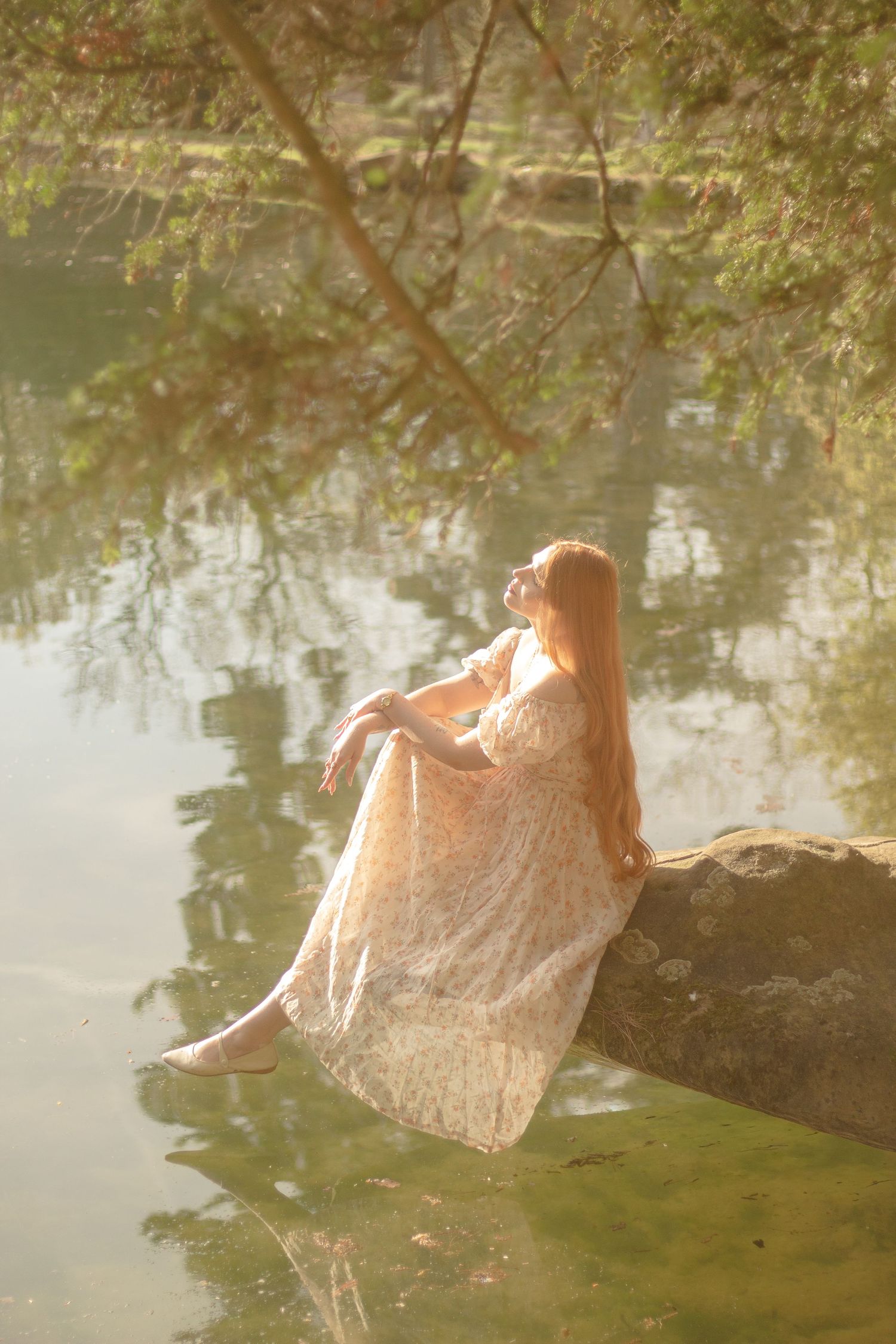 A woman with long red hair is sitting in a rock by a calm body of water, wearing a floral dress.