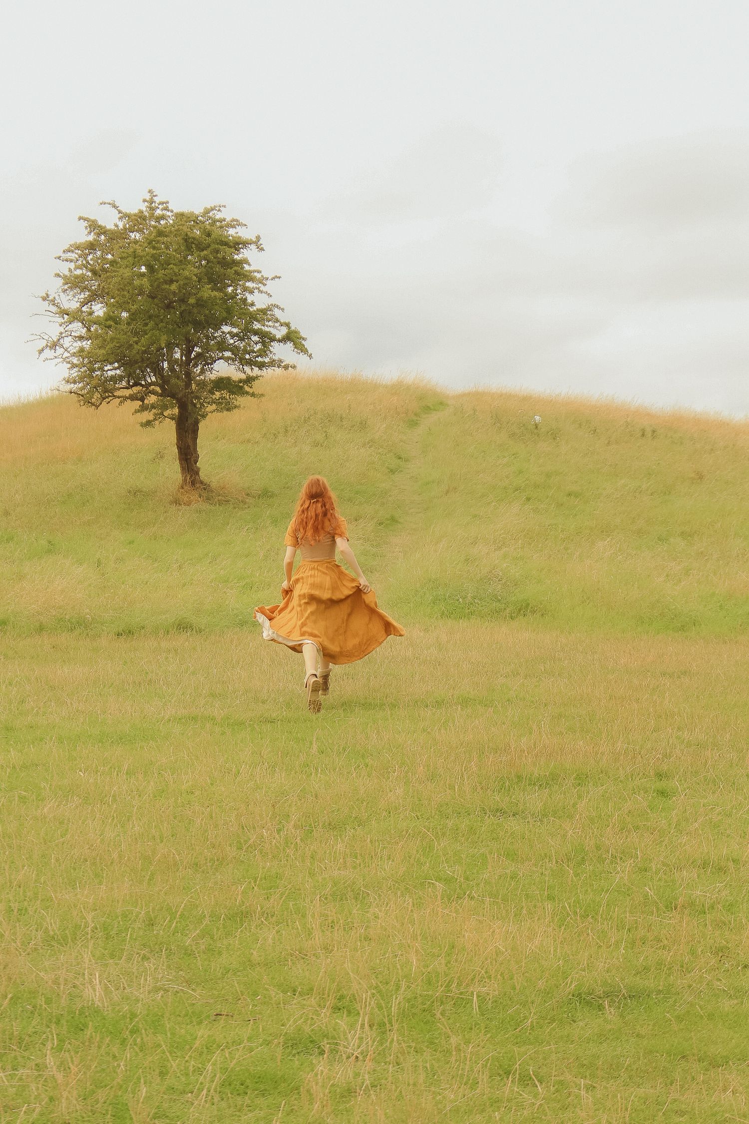 A woman with long ginger hair is running in a field with a single tree in sight, she is wearing a long mustard dress and a corset.