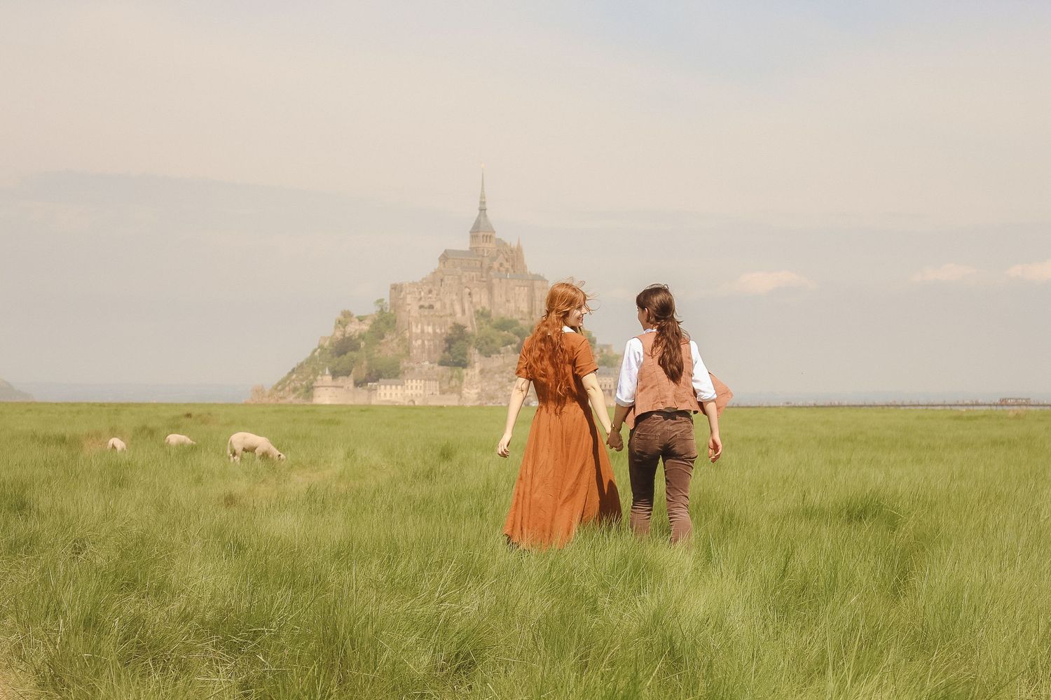 A couple are hand in hand standing in a field with sheeps and in the background you can see the Mont Saint Michel