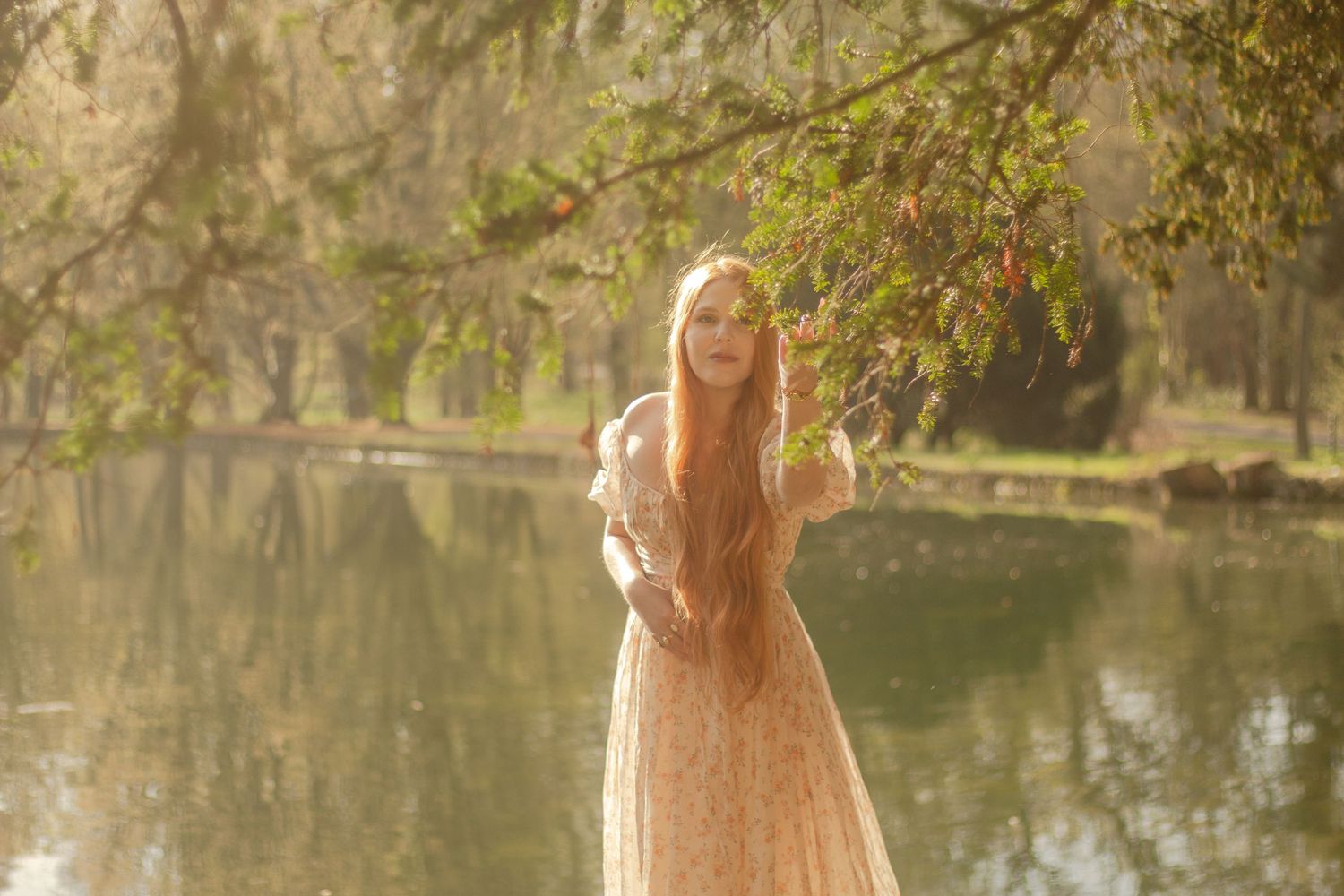 A woman with long red hair is standing by a calm body of water, wearing a floral dress.