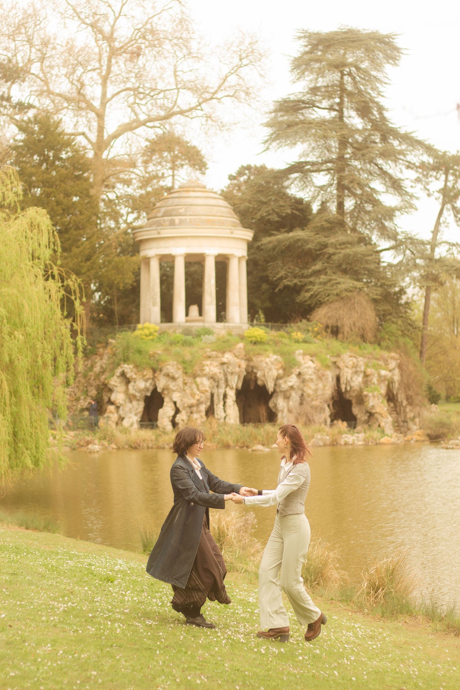 A lesbian couple is running to an embrace, in the background there is a willow tree and a temple located in Bois de Vincennes.