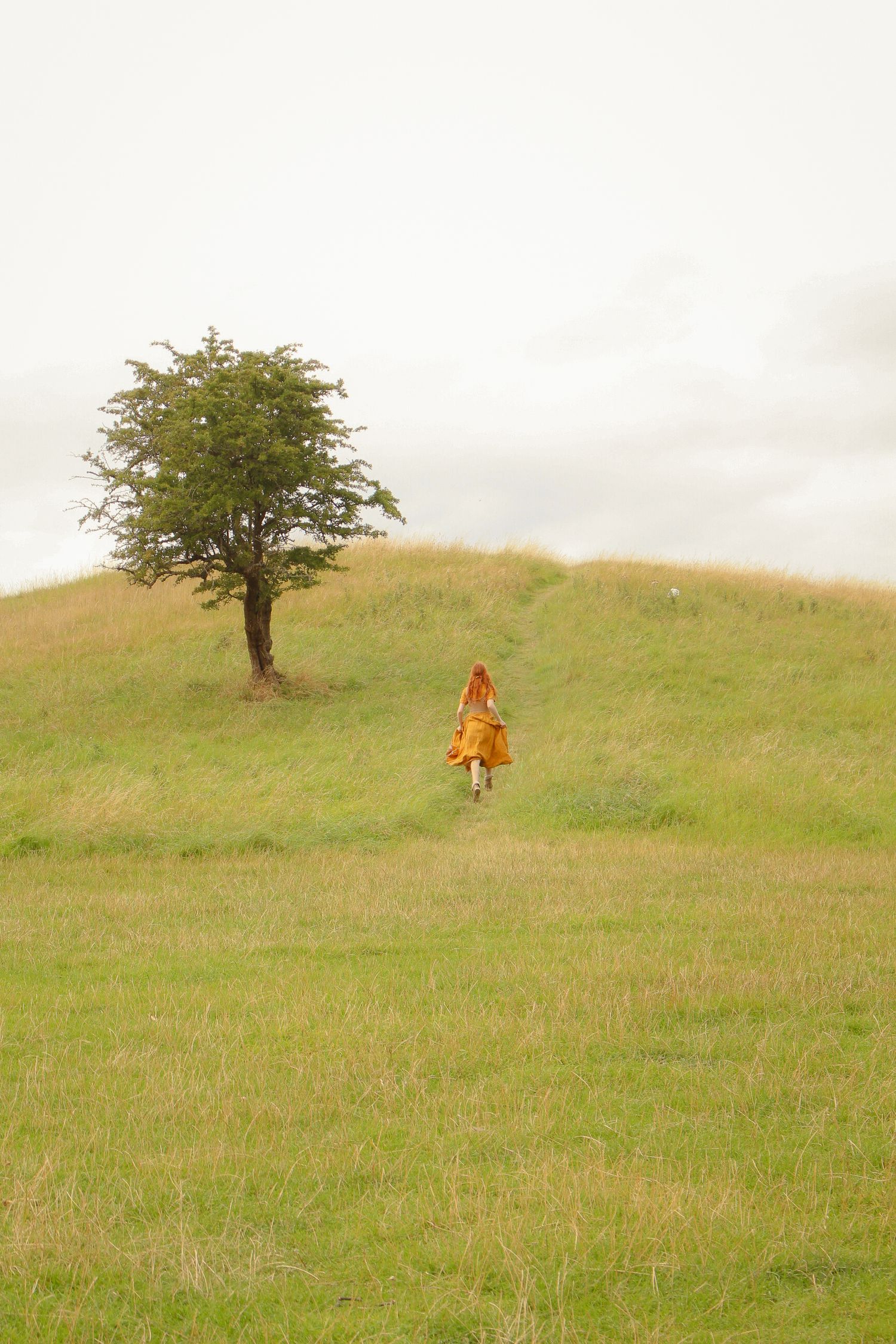 A woman with long ginger hair is running in a field with a single tree in sight, she is wearing a long mustard dress and a corset.
