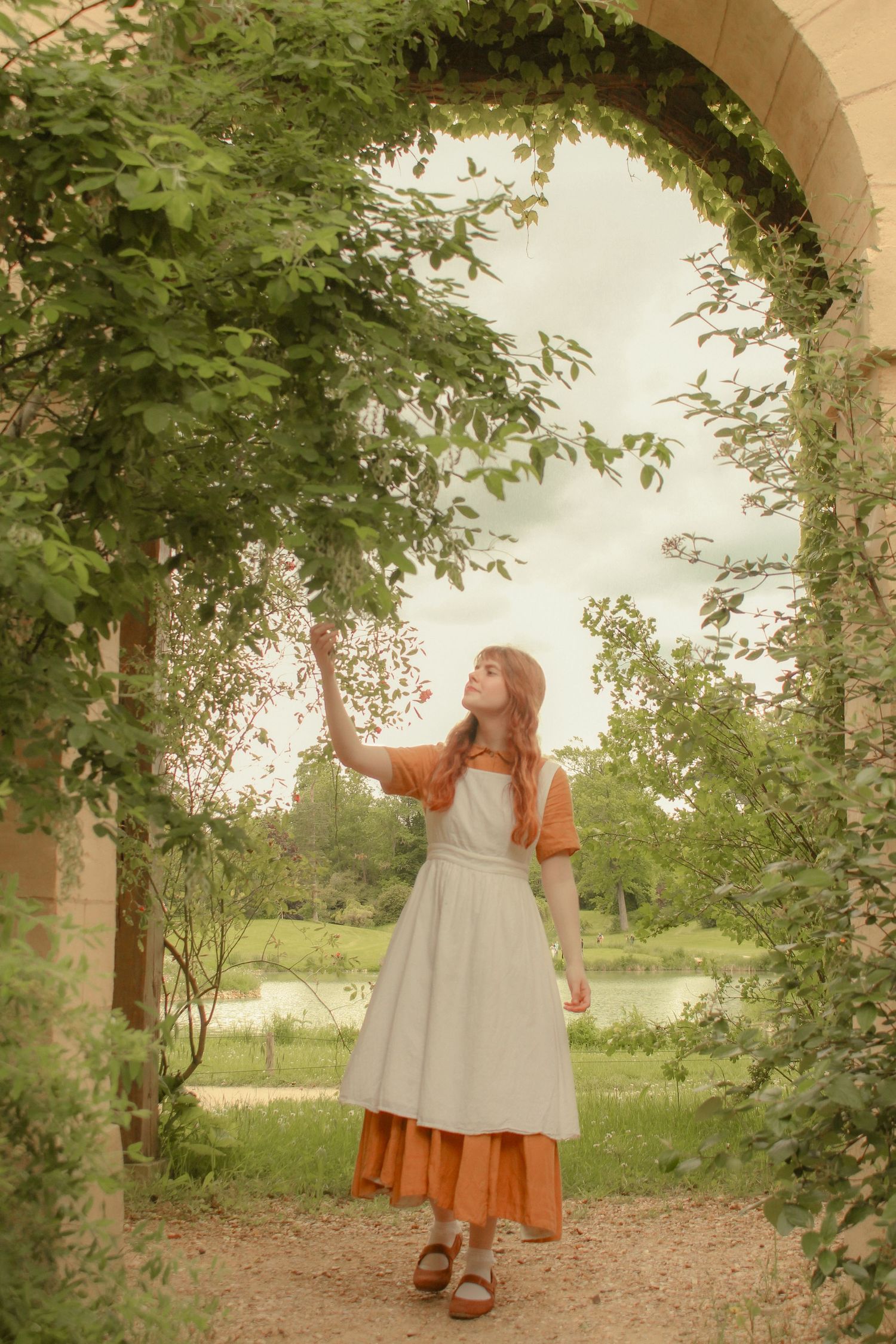 A woman with long red hair is standing near an arch and touching a branch of a plant, she is wearing a long dress with an apron.