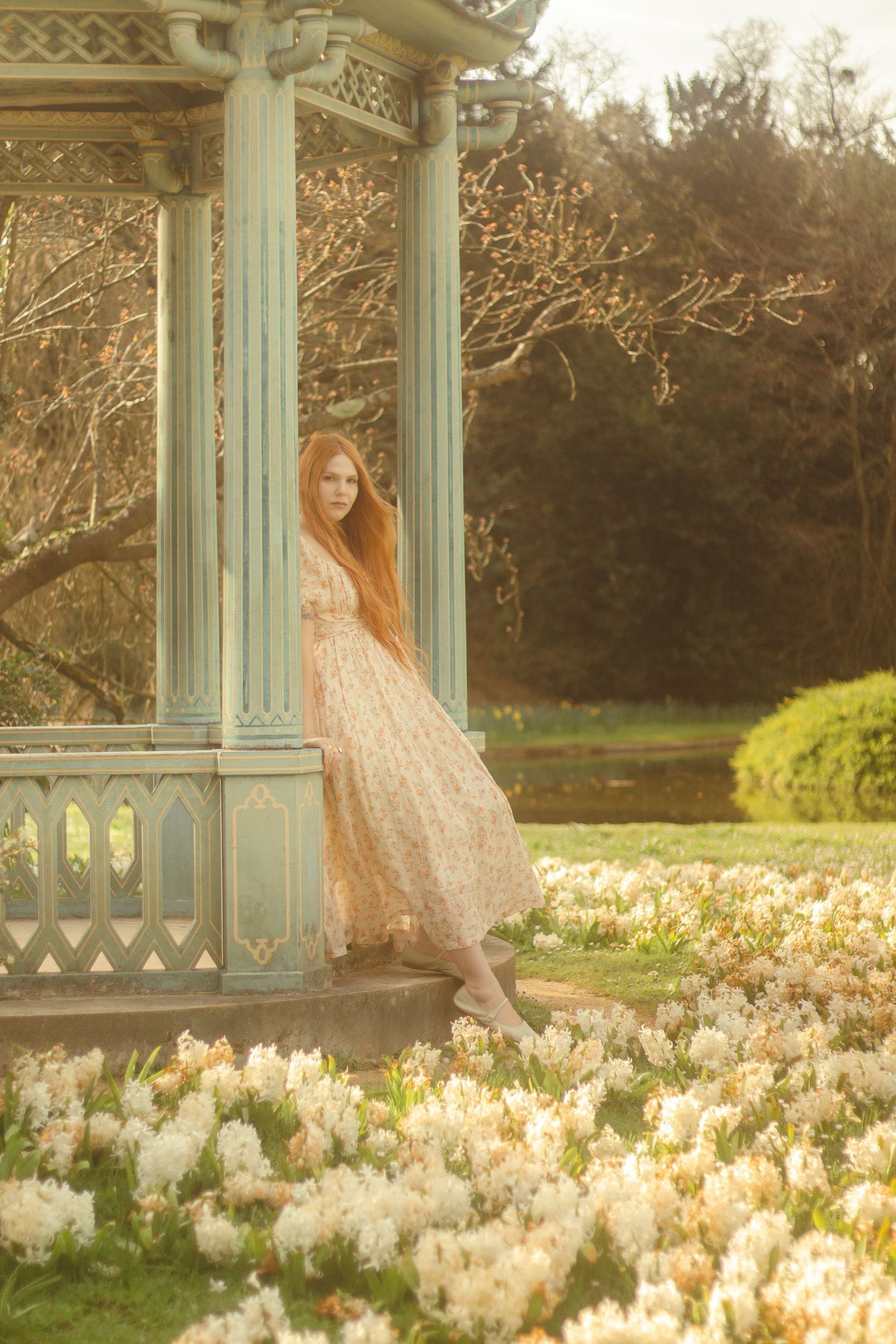 A woman with long red hair is standing by a decorative gazebo, wearing a floral dress. She is surrounded by a garden filled with flowers.