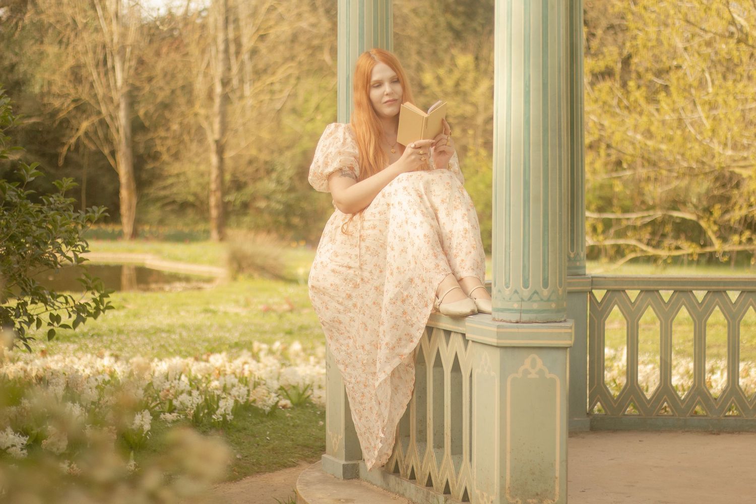 A woman with long red hair wearing a floral dress is sitting and reading a book. There are flowers in the background