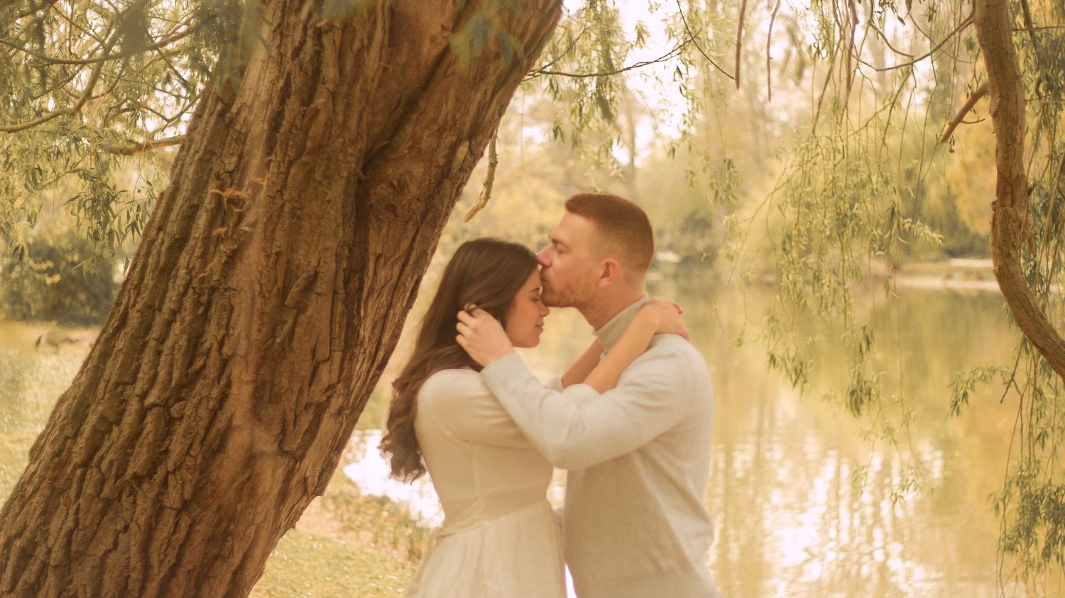 A men is kissing the forehead of her girlfriend underneath a willow tree, she has her arms around his neck, the light of the photo is soft.