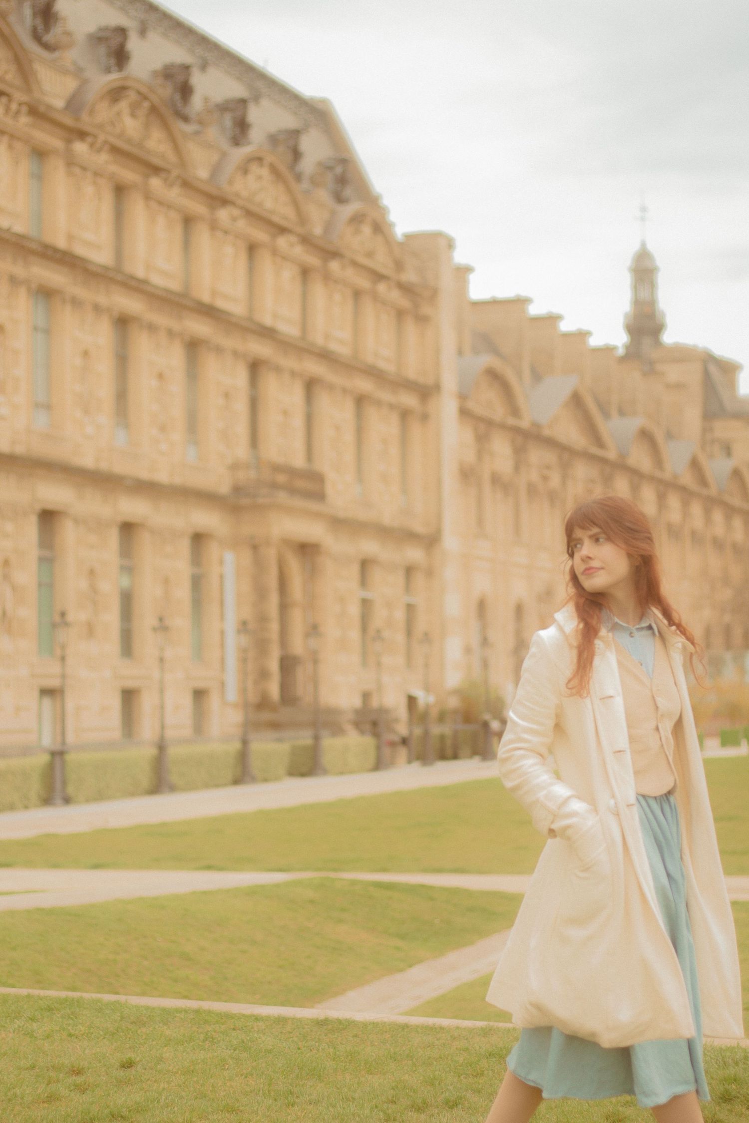 A woman with long auburn hair is walking in Paris, wearing a vintage outfit. In the background there is the buildings near the Louvre.