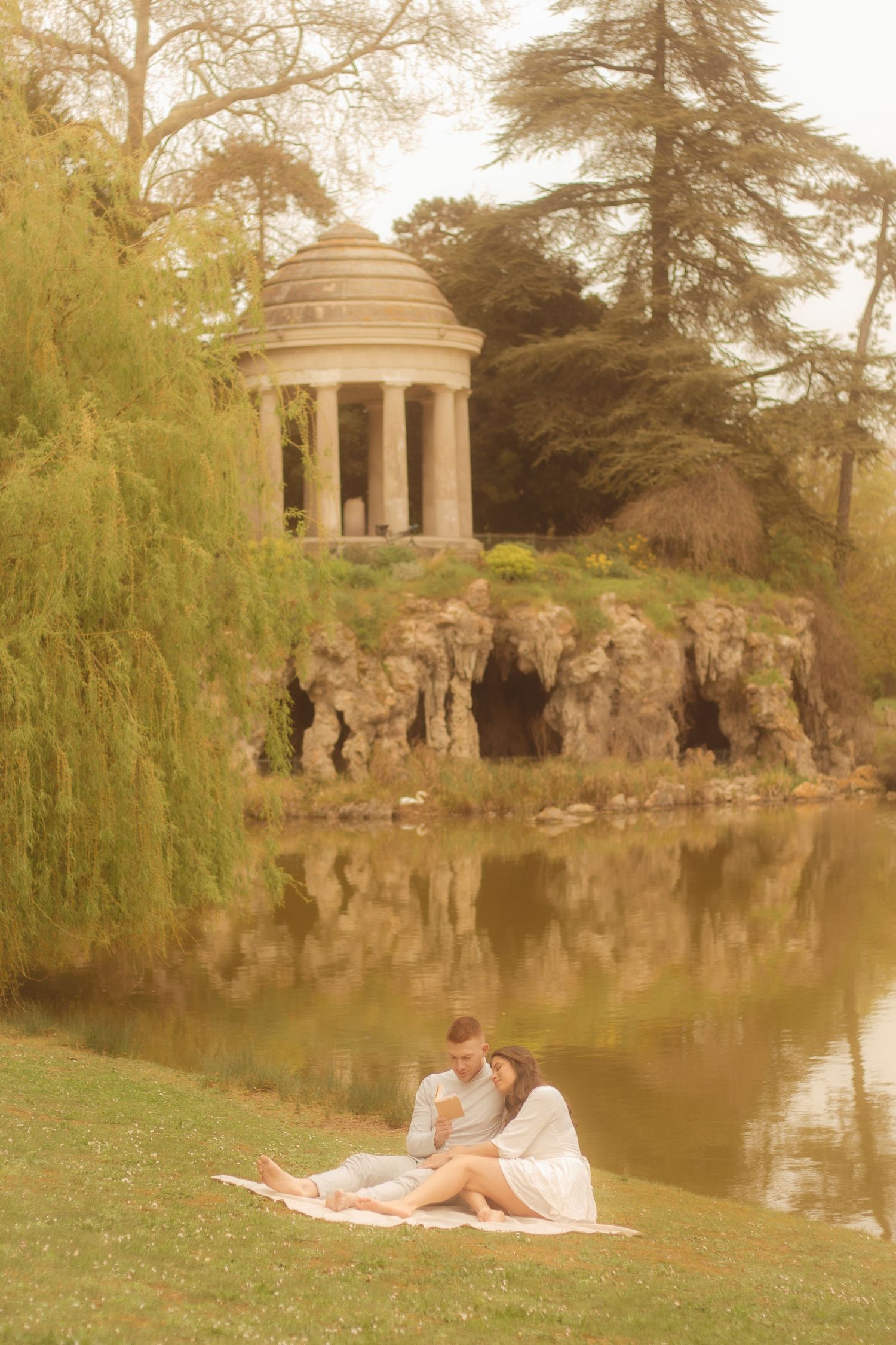 A couple is sitting in the grass reading a book, in the background there is a willow tree and a temple located in Bois de Vincennes.