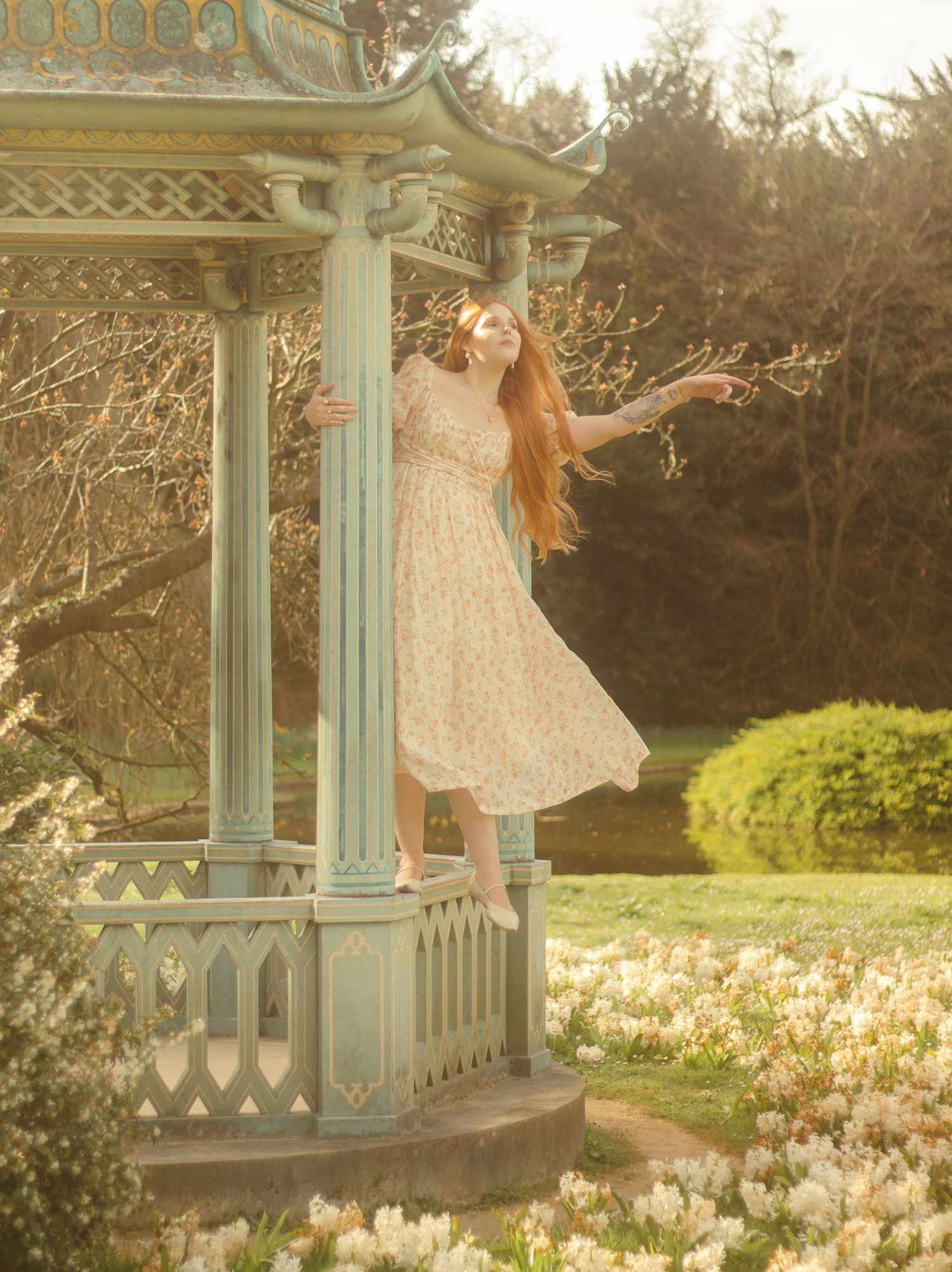 A woman with long red hair is standing in a decorative gazebo, wearing a floral dress. She is surrounded by a garden filled with flowers.