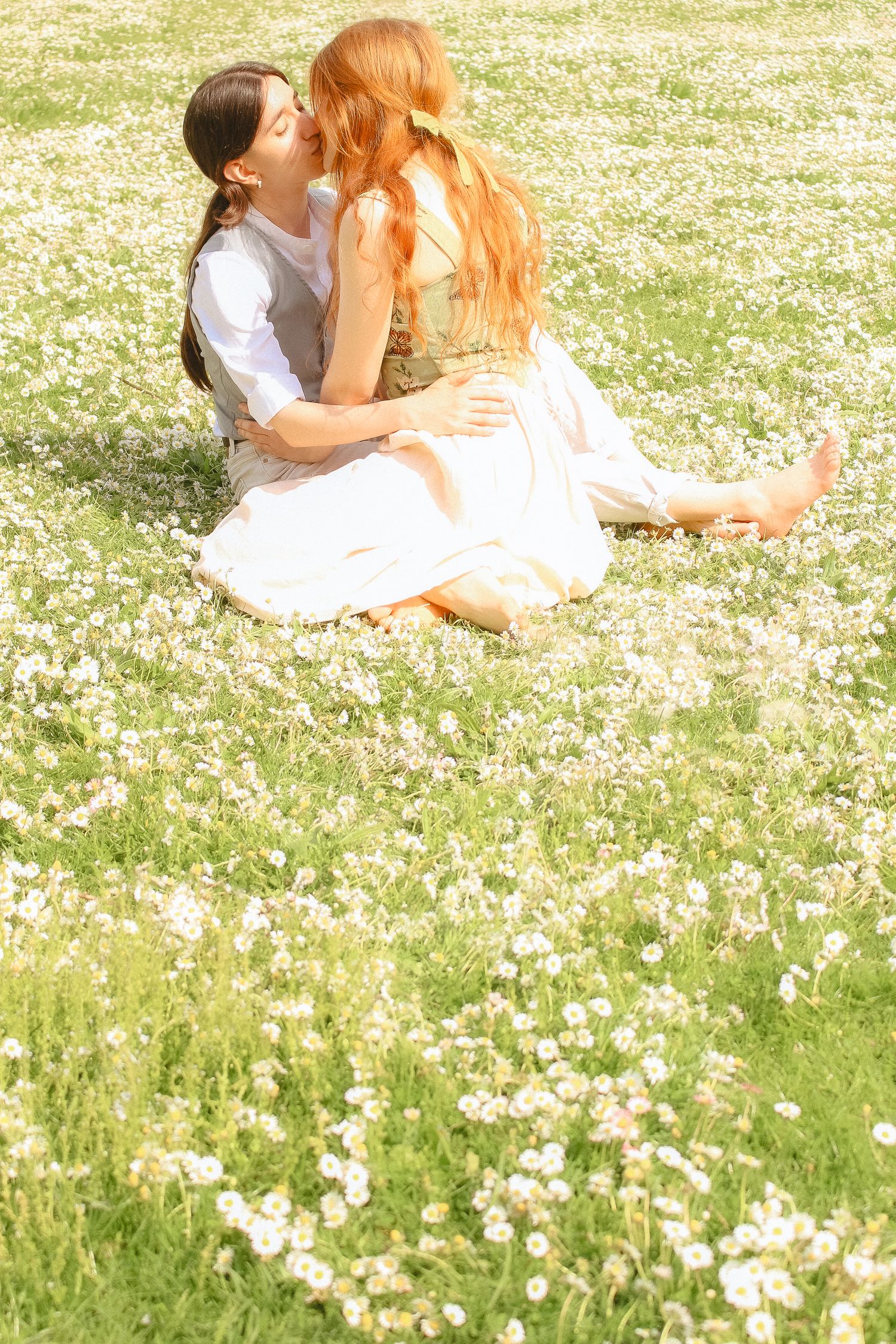 A couple is kissing in a field of tiny daises in spring, the girl on top has a bow in her hair.