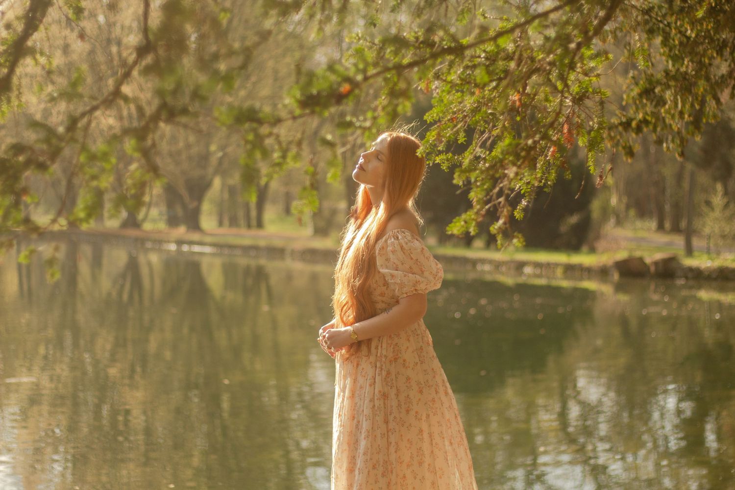 A woman with long red hair is standing by a calm body of water, wearing a floral dress. The light shines on her hair.