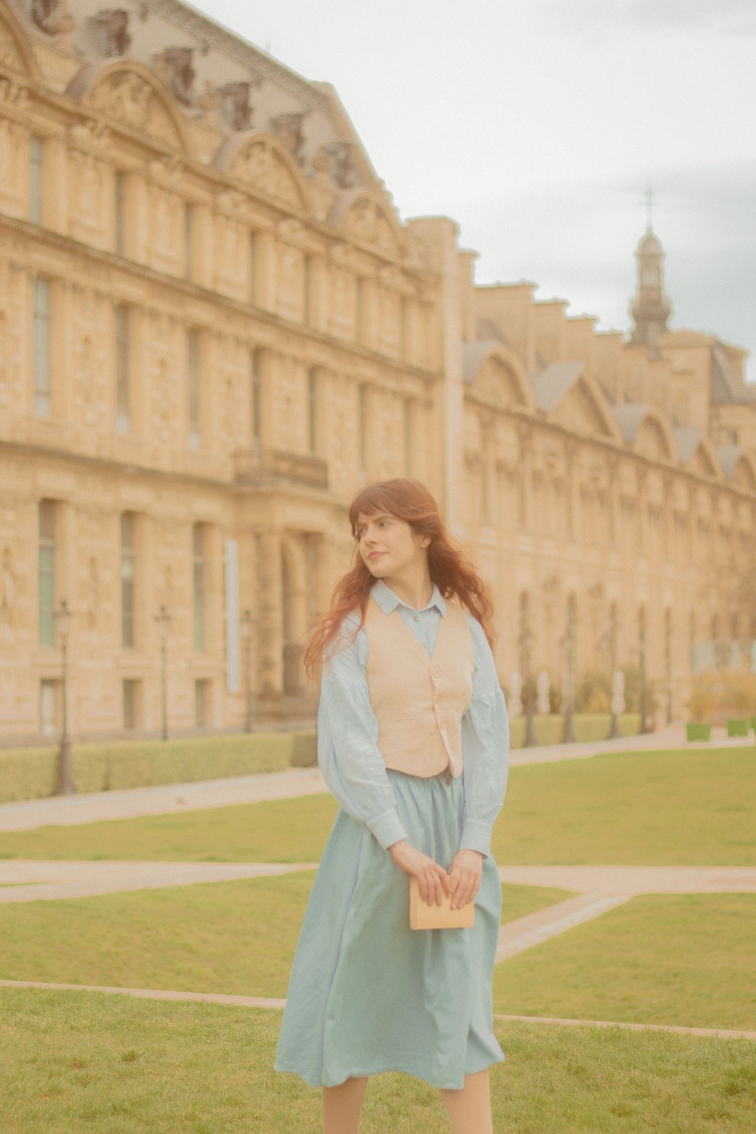 A woman with long auburn hair is walking in Paris, wearing a vintage outfit. In the background there is the buildings near the Louvre.