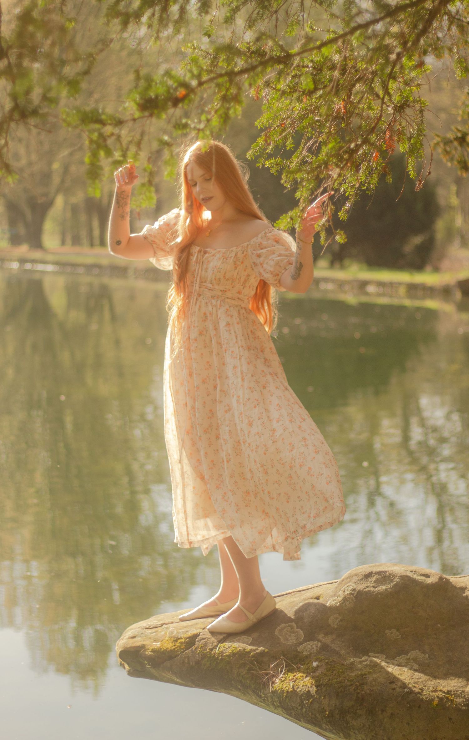 A woman with long red hair is standing by a calm body of water, wearing a floral dress. There is mouvement and light shining.