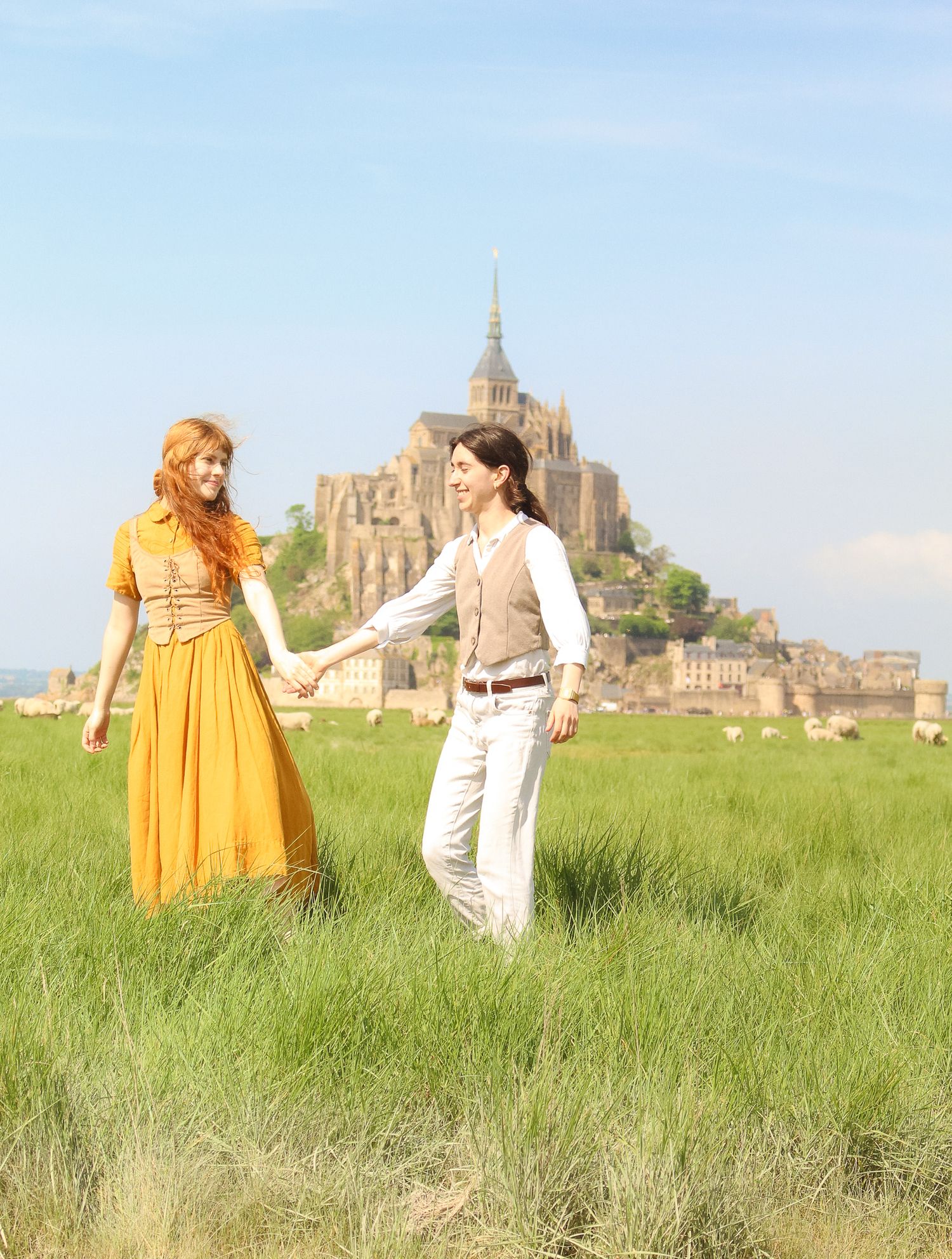 A couple are hand in hand walking in a field with sheeps and in the background you can see the Mont Saint Michel