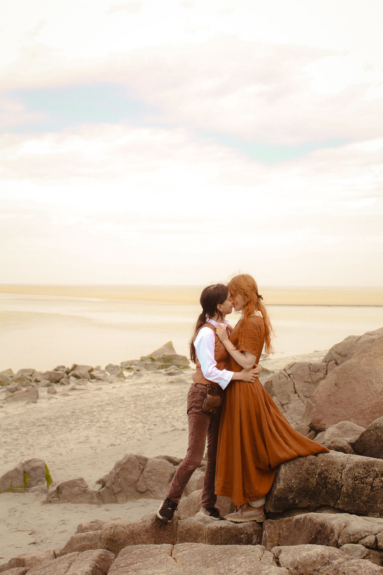 A couple is about to kiss, they are dressed in a vintage style, both are wearing brown. They are located in a beach, standing by the rocks.