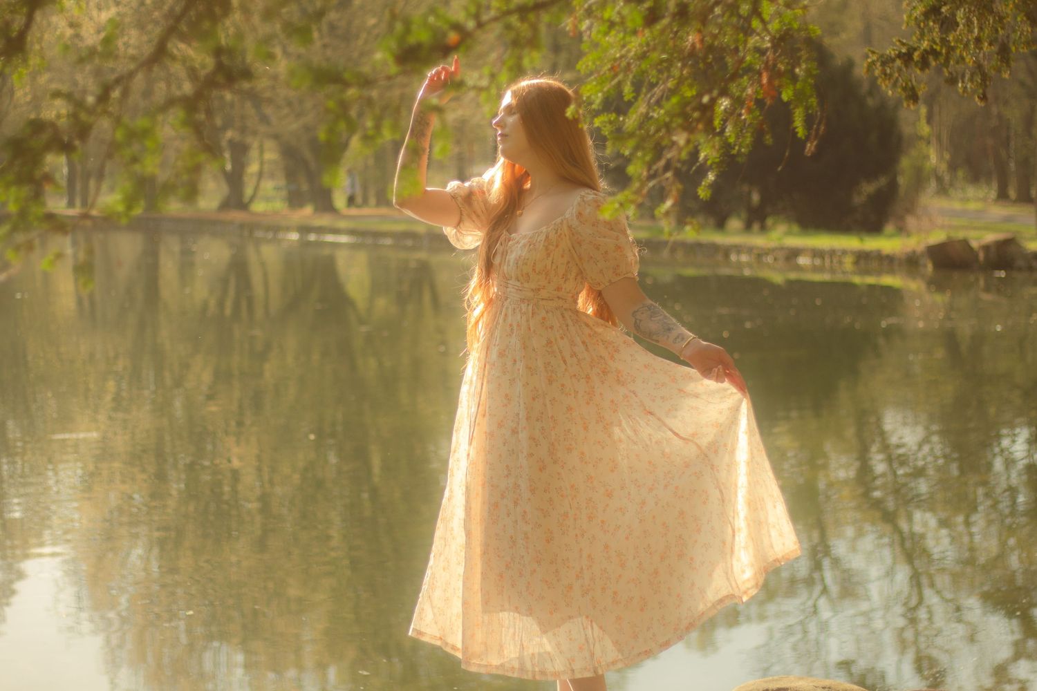 A woman with long red hair is standing in a rock by a calm body of water, wearing a floral dress.