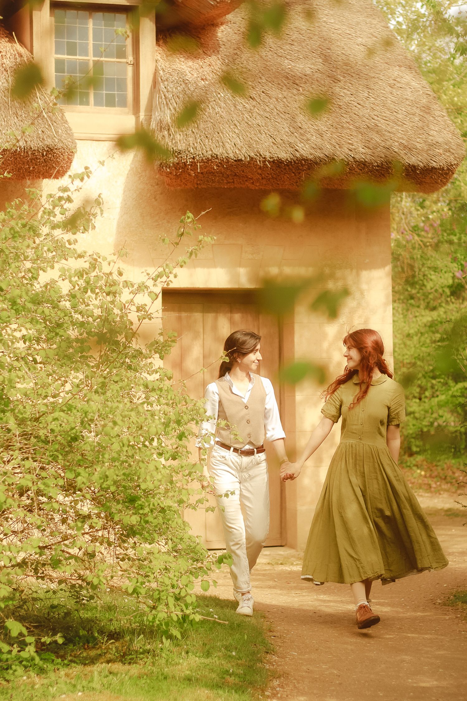 A couple is walking hand in hand surround by plants and near a cottage.