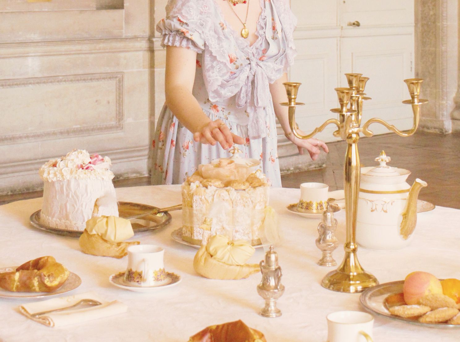 A woman in a floral dress leans to grab a piece of a sweet treat on a table full of cakes and tea cups.