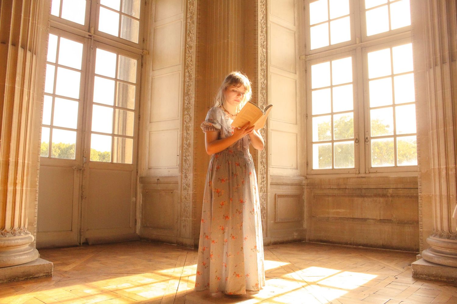 A girl is standing in a piece with long glass windows inside a baroque castle in Paris, she is reading a book and has a long dress.