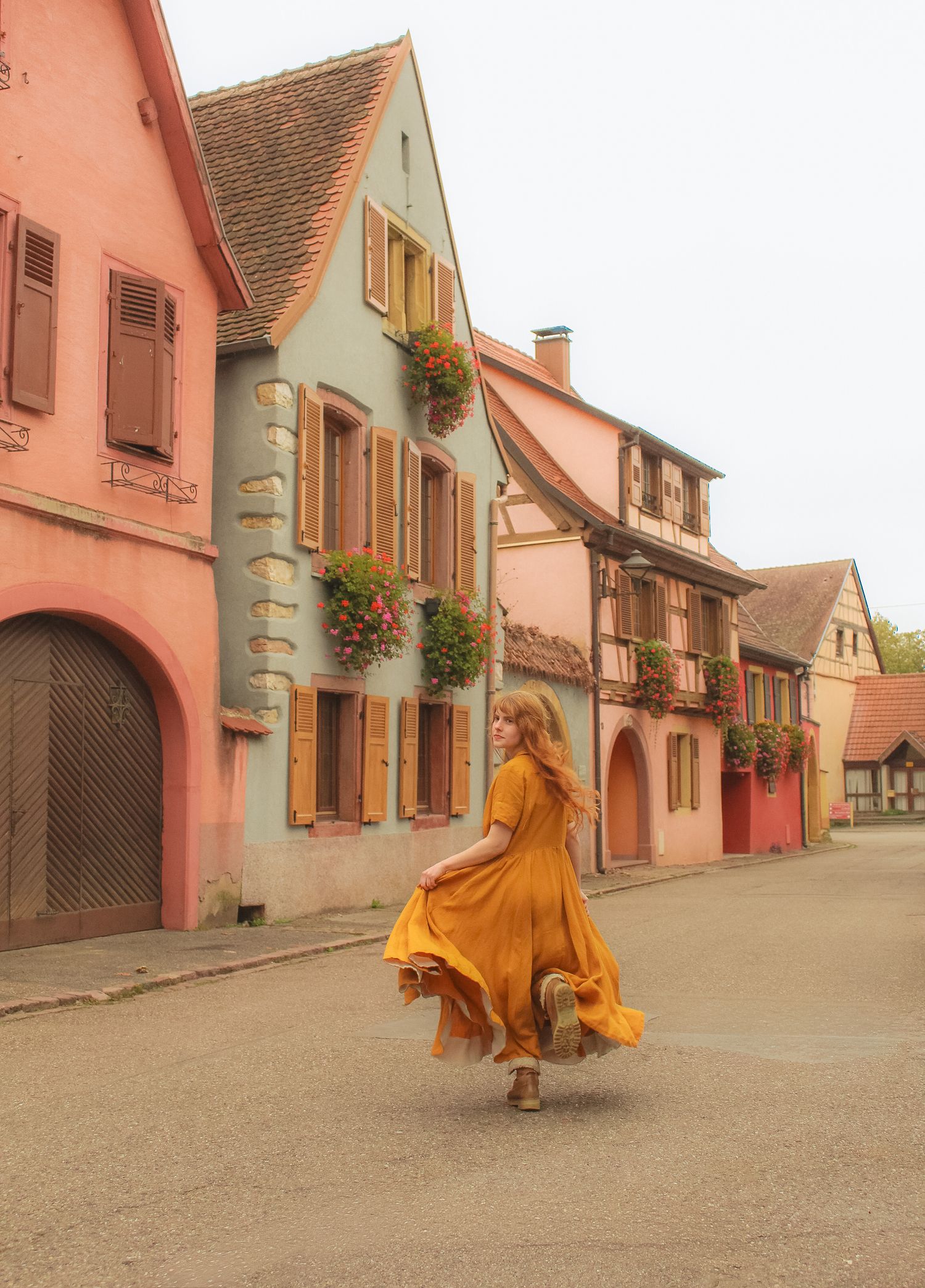 A girl is running looking behind her,  she is wearing a long flowy dress and coat a book. There are colored houses in the background