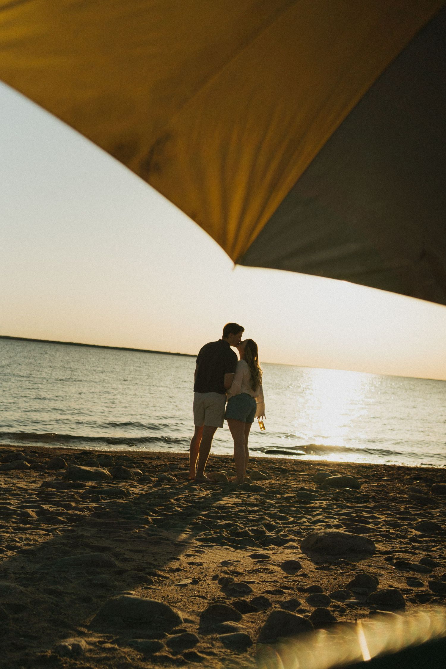 A couple shares a kiss under a beach umbrella in the sunset.