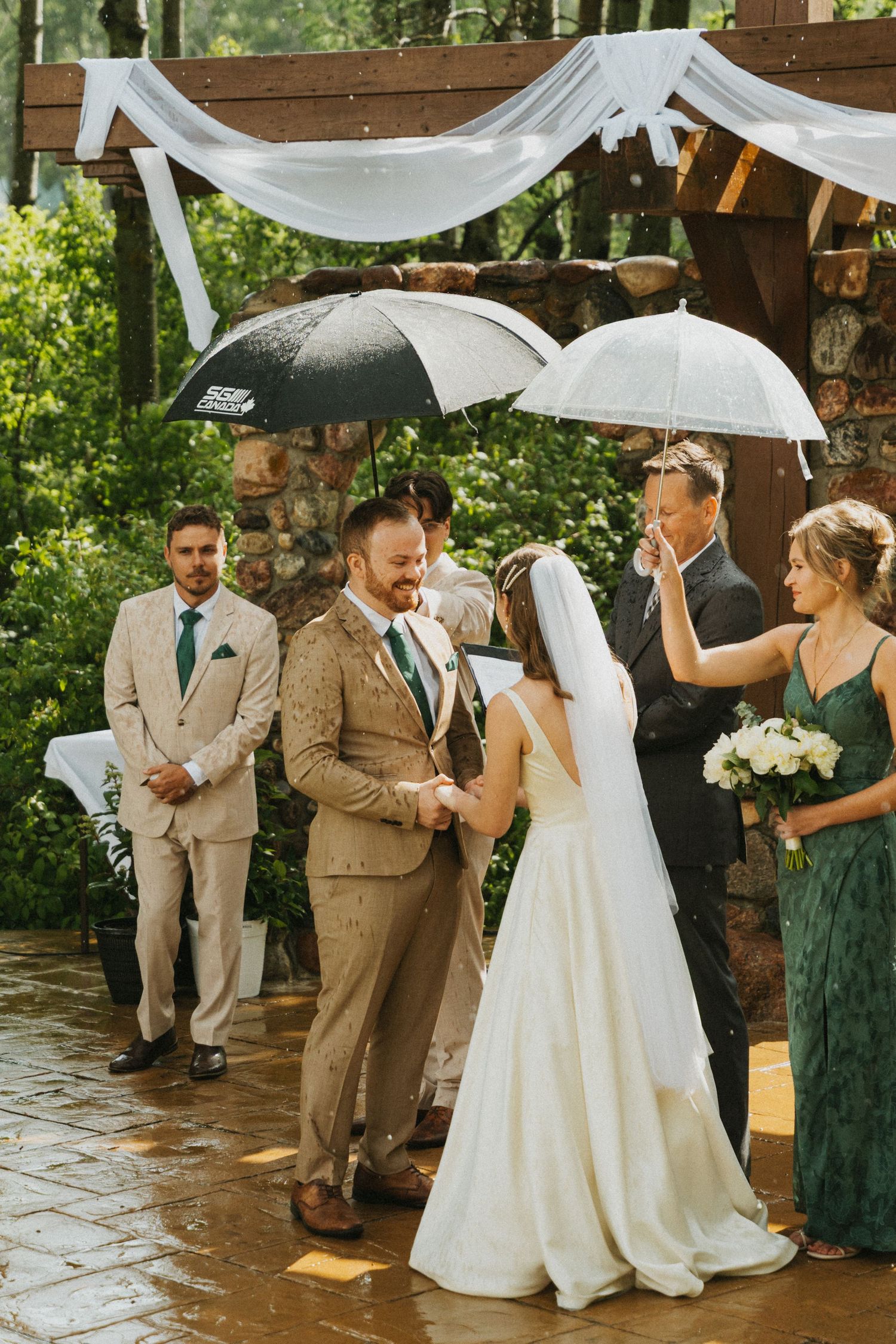 A candid photo of a couple exchanging vows on their wedding day as it rain on them. They hold umbrellas and laugh.