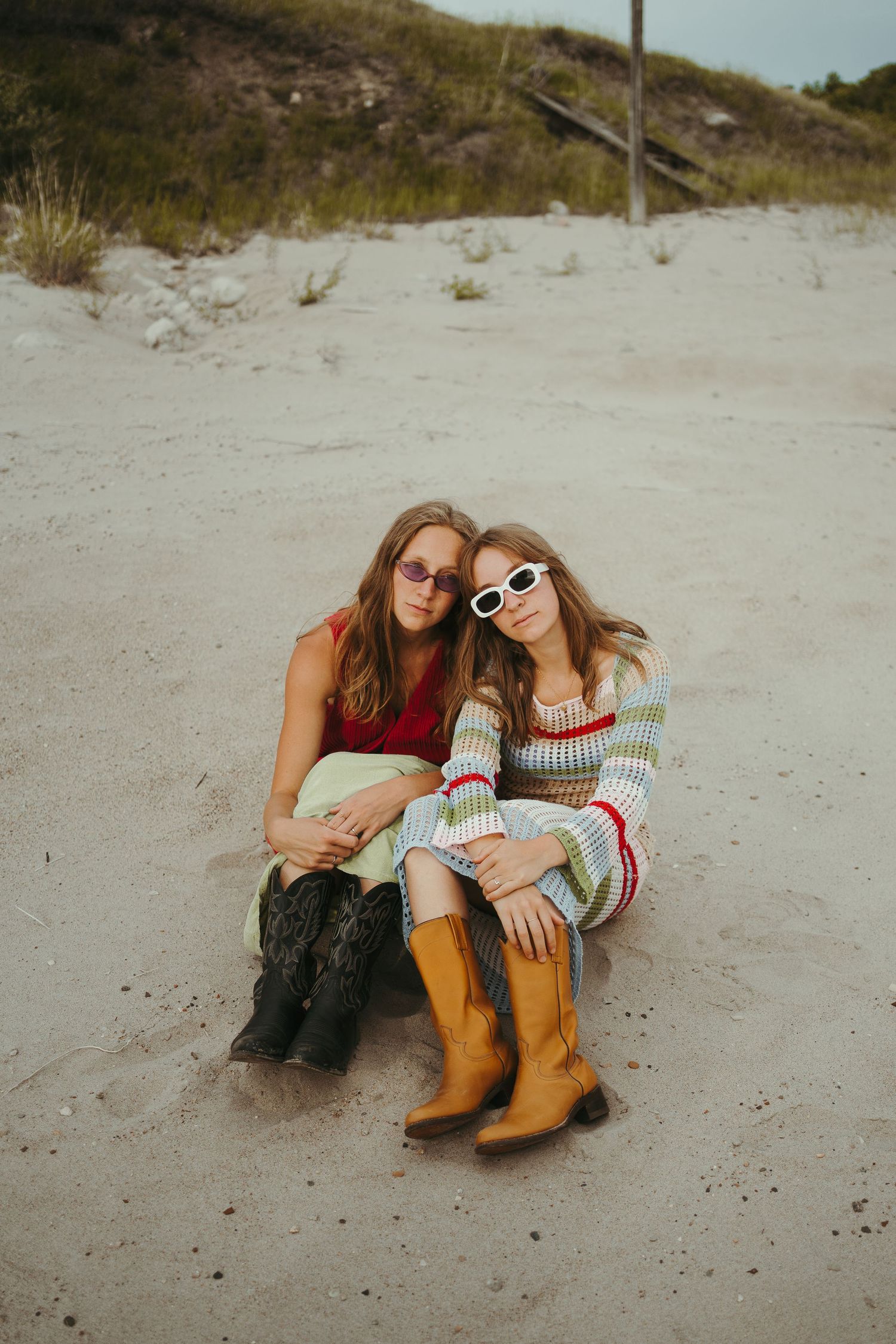 A couple of girls sit in the sand wearing funky outfits