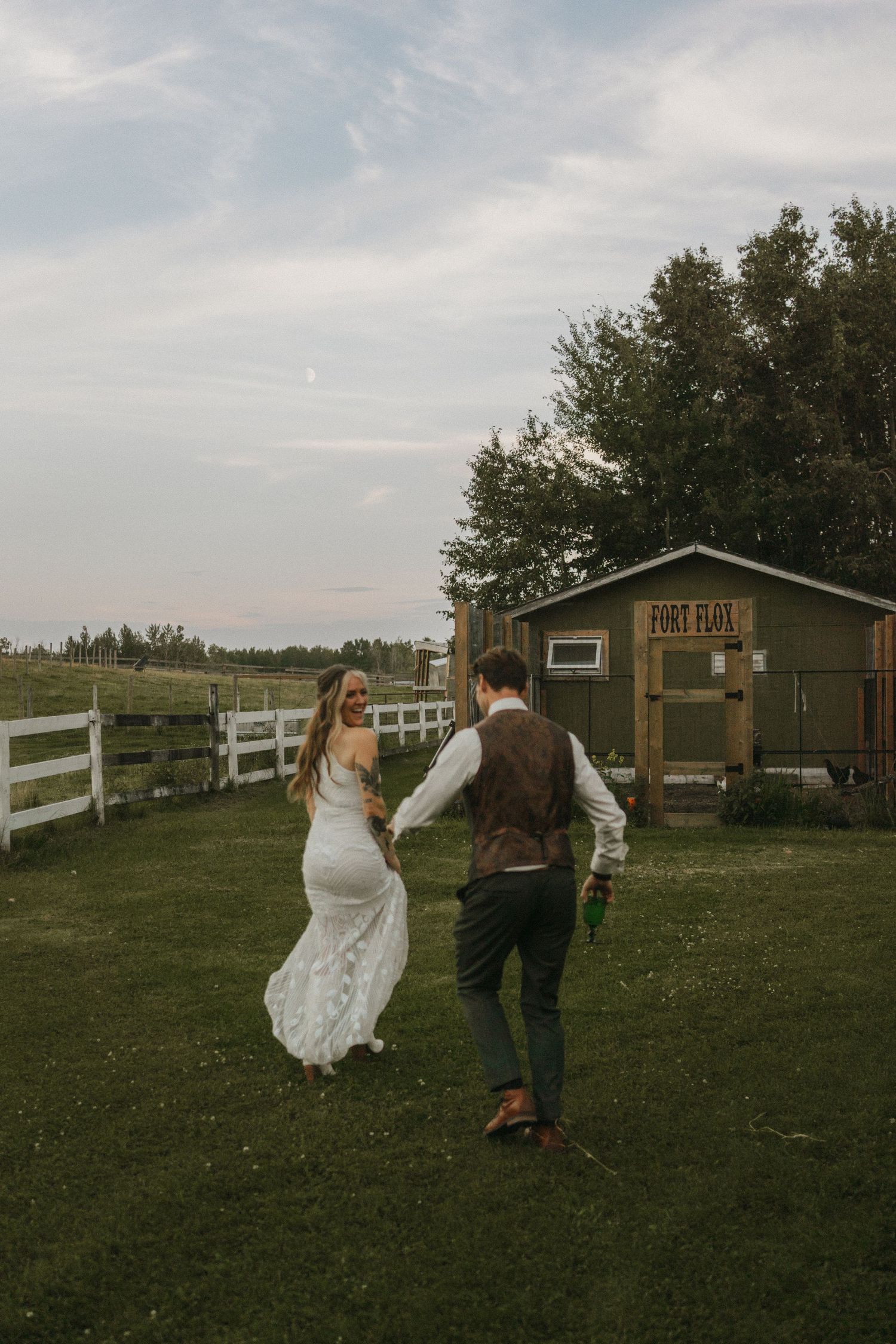 A photo of a bride and groom running around on their farm after getting married. The man chases the woman as they laugh.