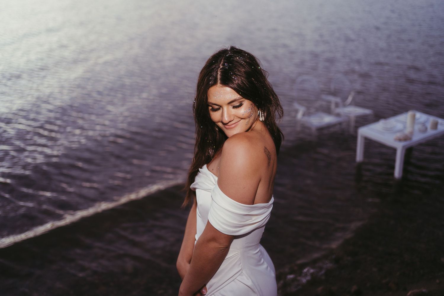 A bride at the ocean. She has sparkles on her faces and the water is purple