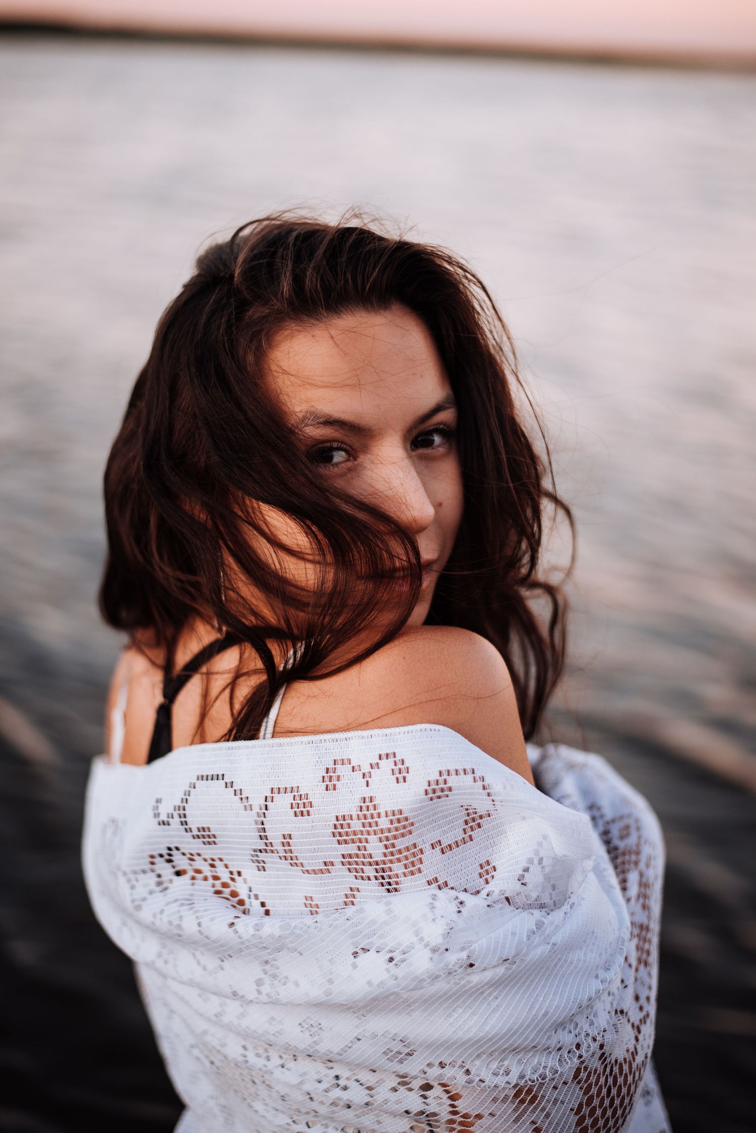 A woman stares into the camera at the lake. She is covered in lace and the wind blows her hair