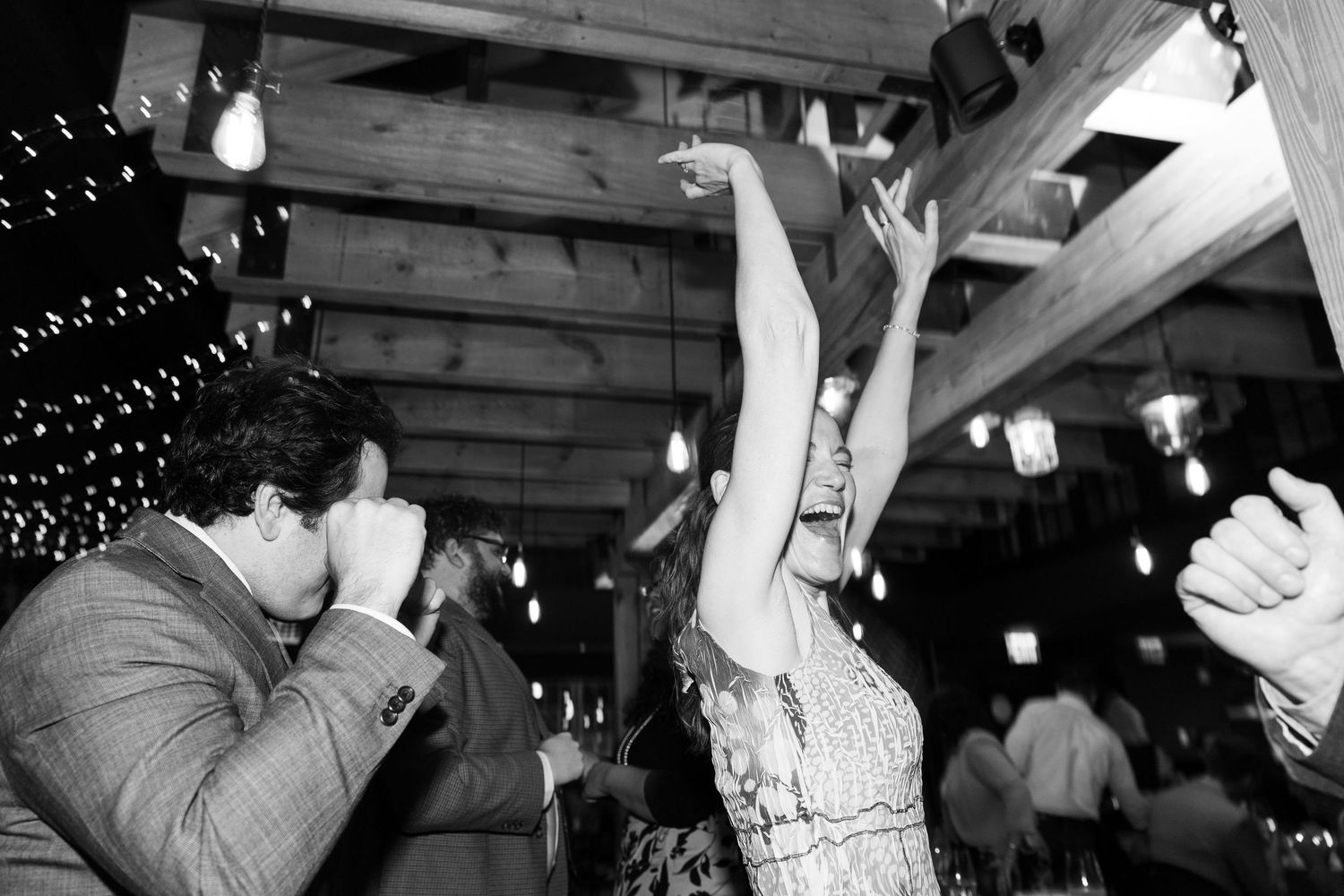 Black and white photo of people dancing and celebrating at a party under string lights and wooden beams.