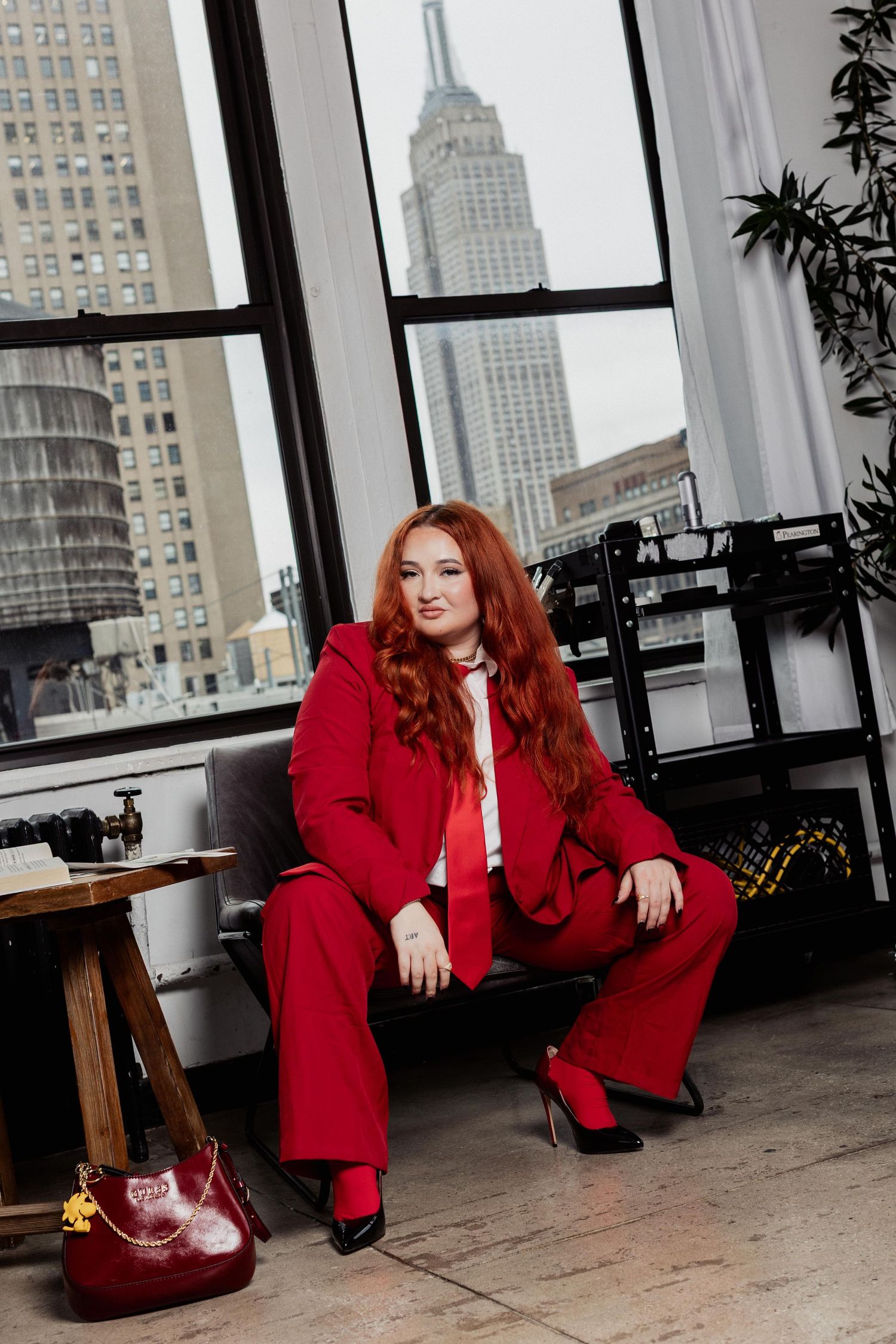 A woman in a red suit sits by a window with the Empire State Building visible in the background.
