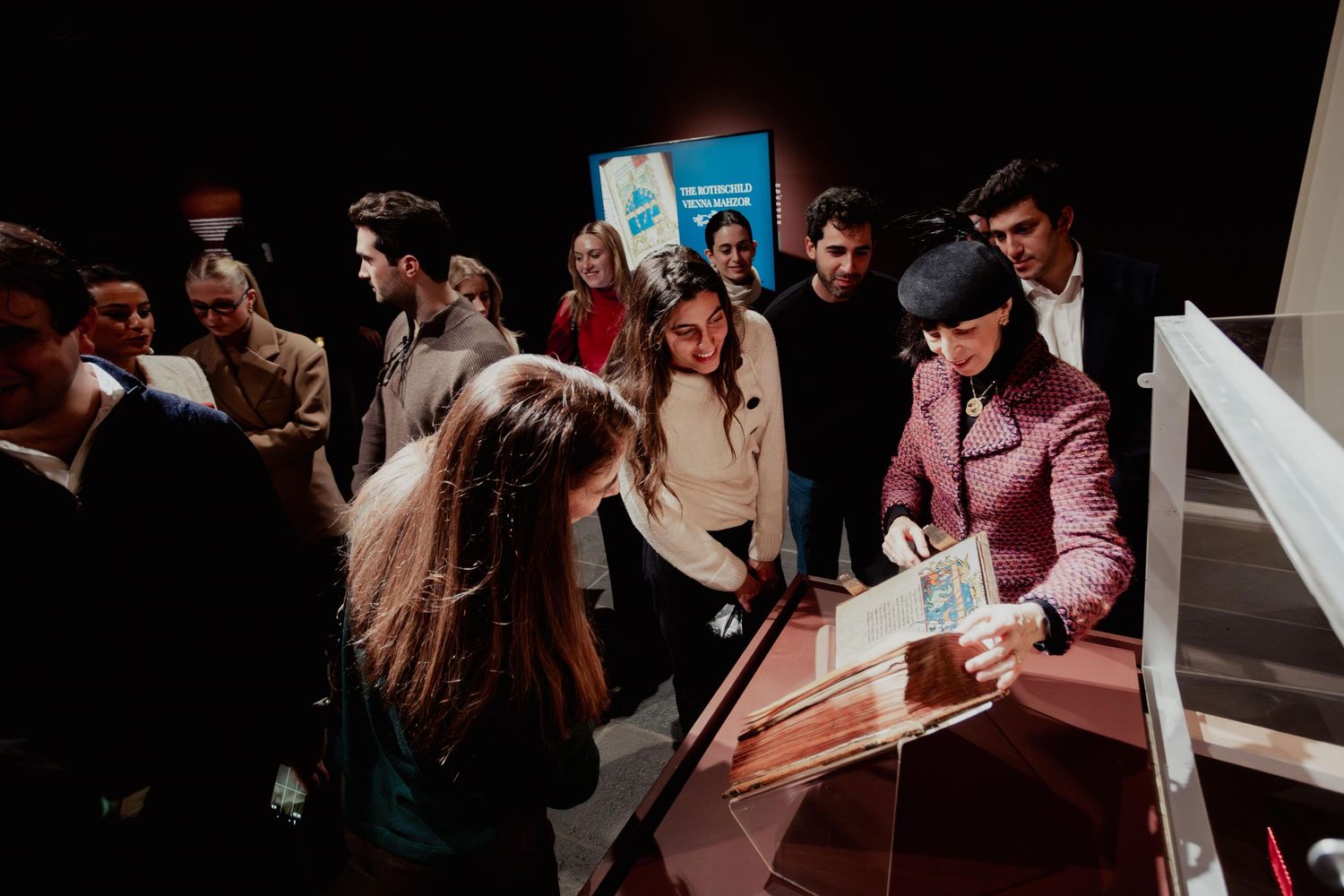 A group of people gather around a white display table in a dimly lit room during an event or exhibition.