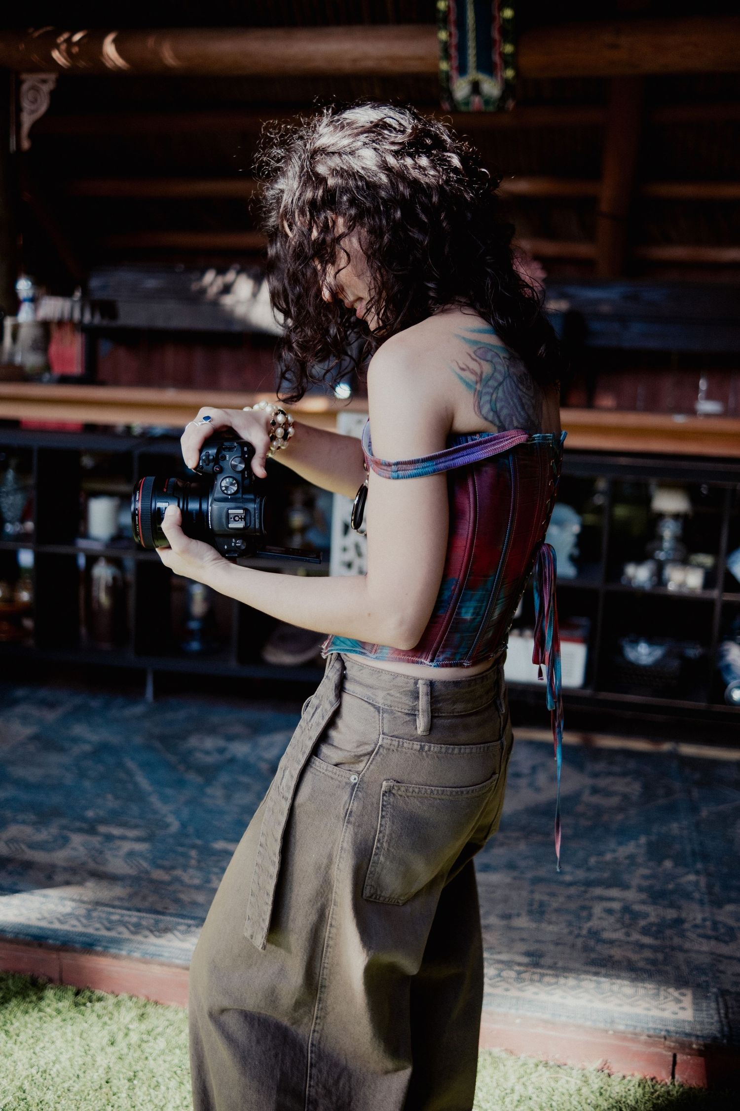 Young woman with tattoos holding a DSLR camera, wearing a plaid crop top and wide-leg pants in a rustic bar setting.