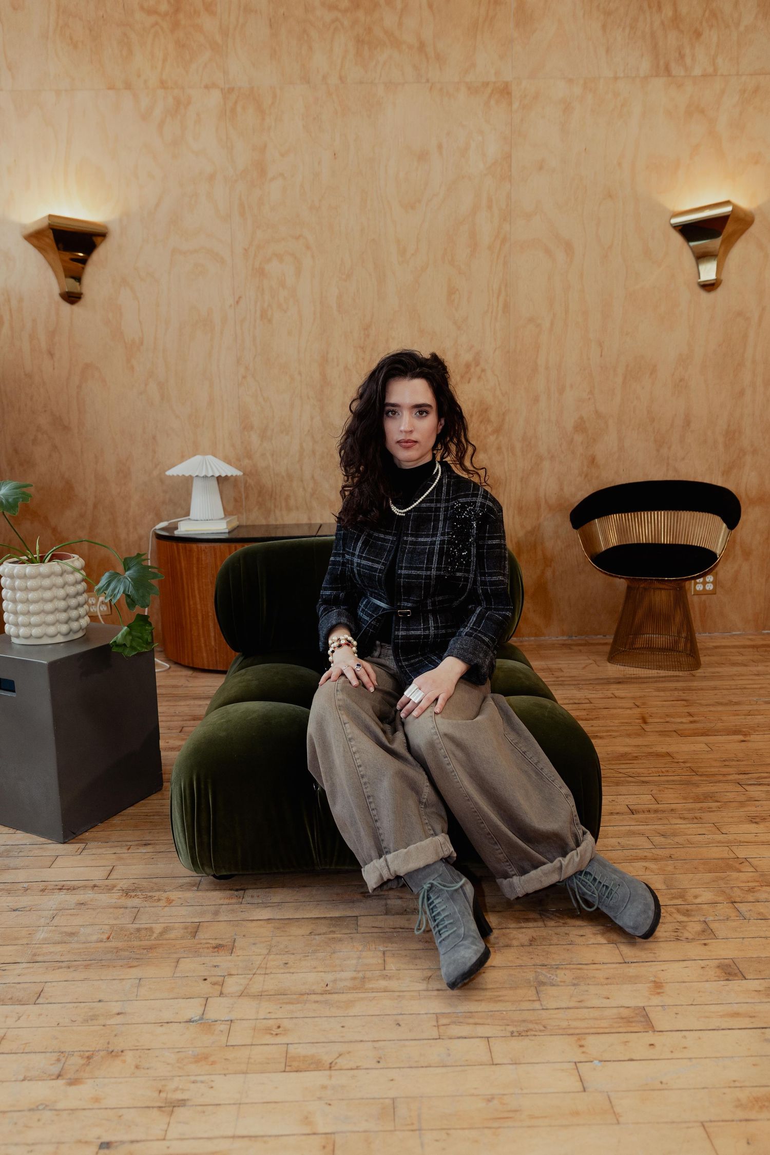 Person wearing plaid jacket and loose pants sits casually on wooden floor in minimalist interior space.