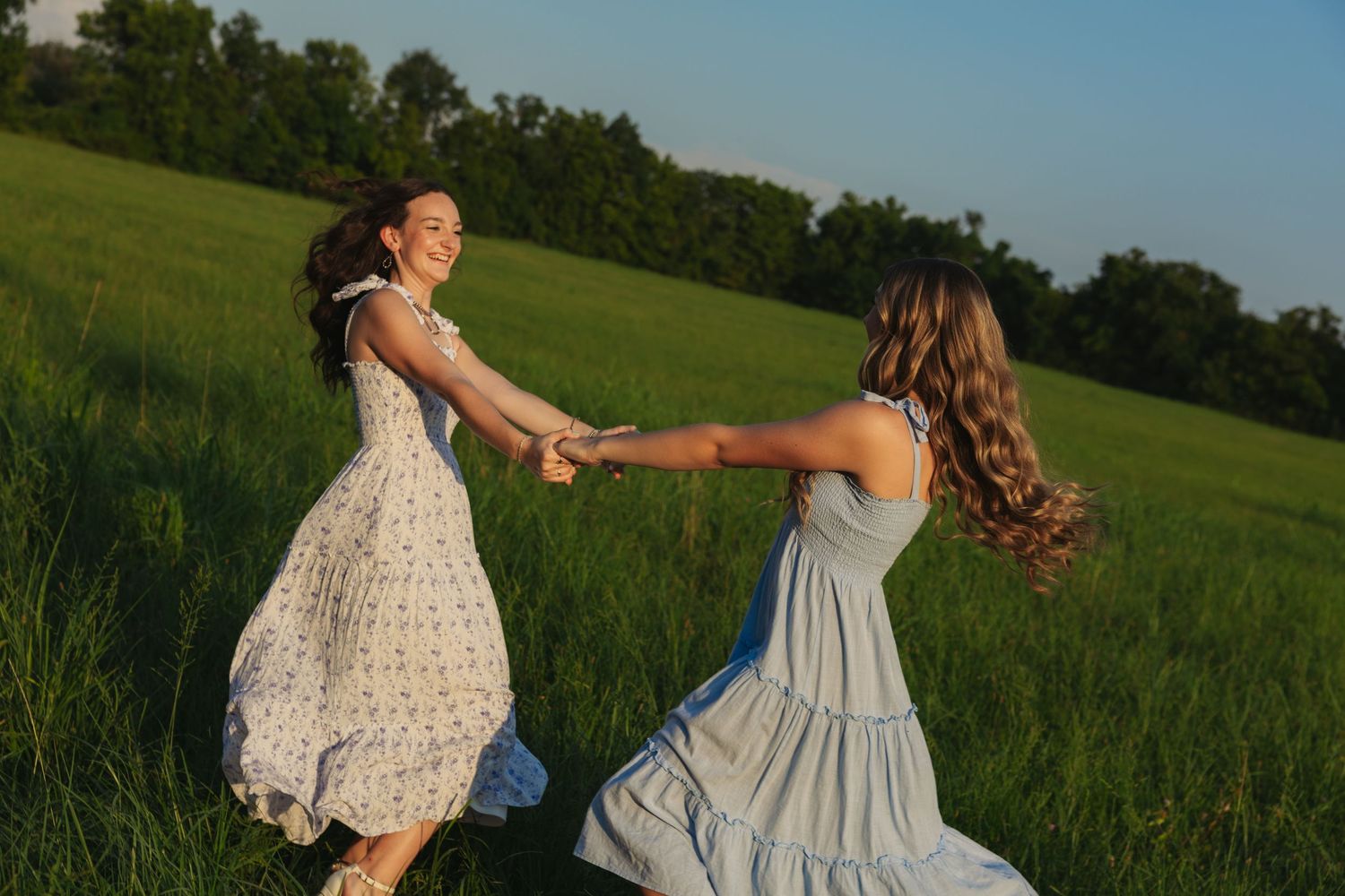 Two young women in summer dresses spinning and laughing together in a lush green field at sunset.