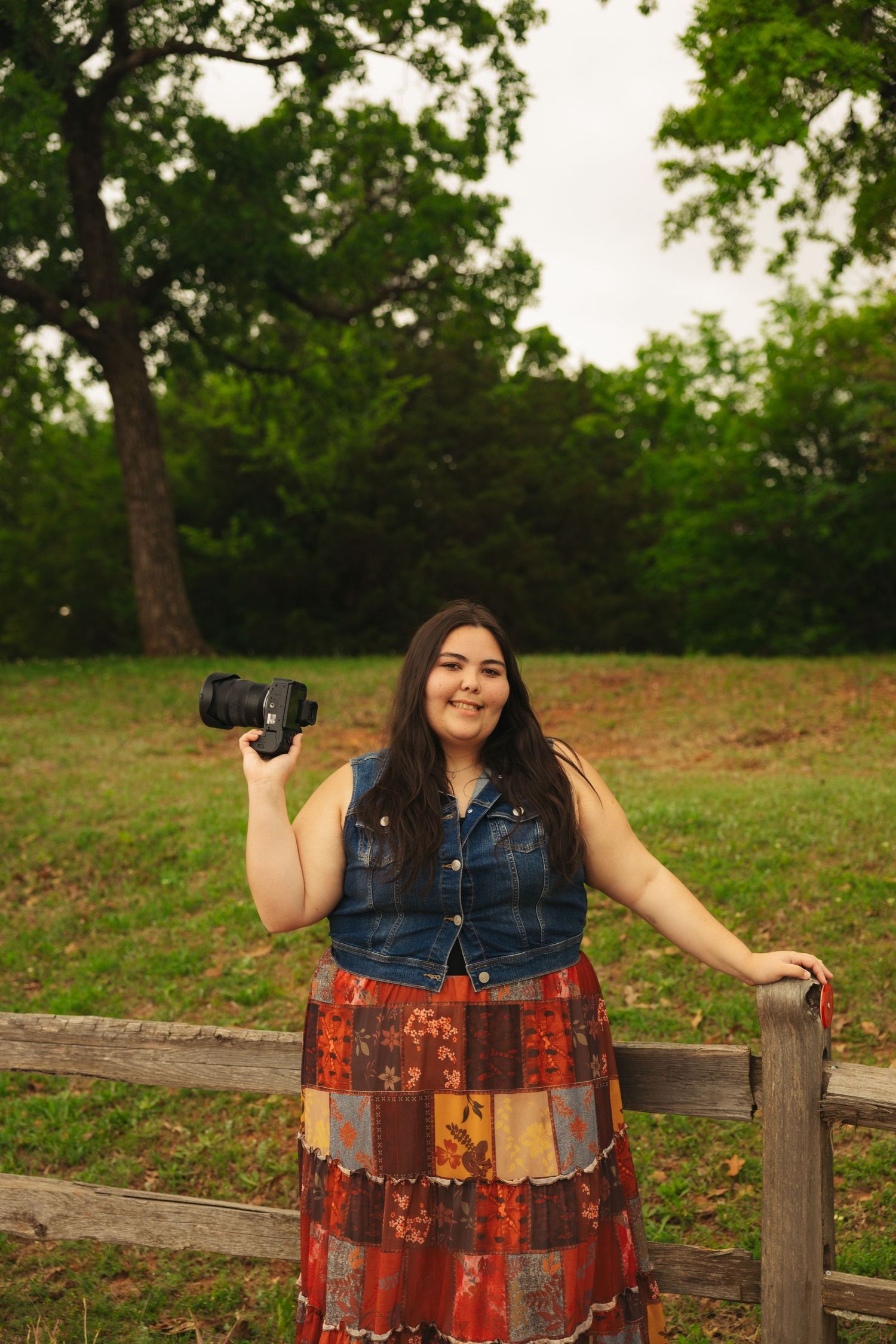 Woman in denim vest and patchwork skirt holds camera while leaning on wooden fence in lush green park.