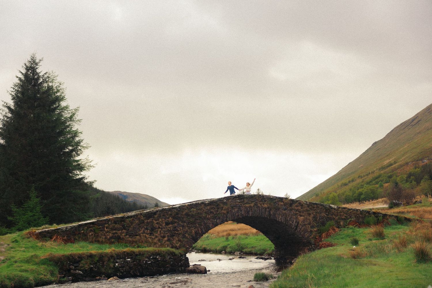 Couple crossing rustic stone arch bridge over stream in misty Scottish Highlands.