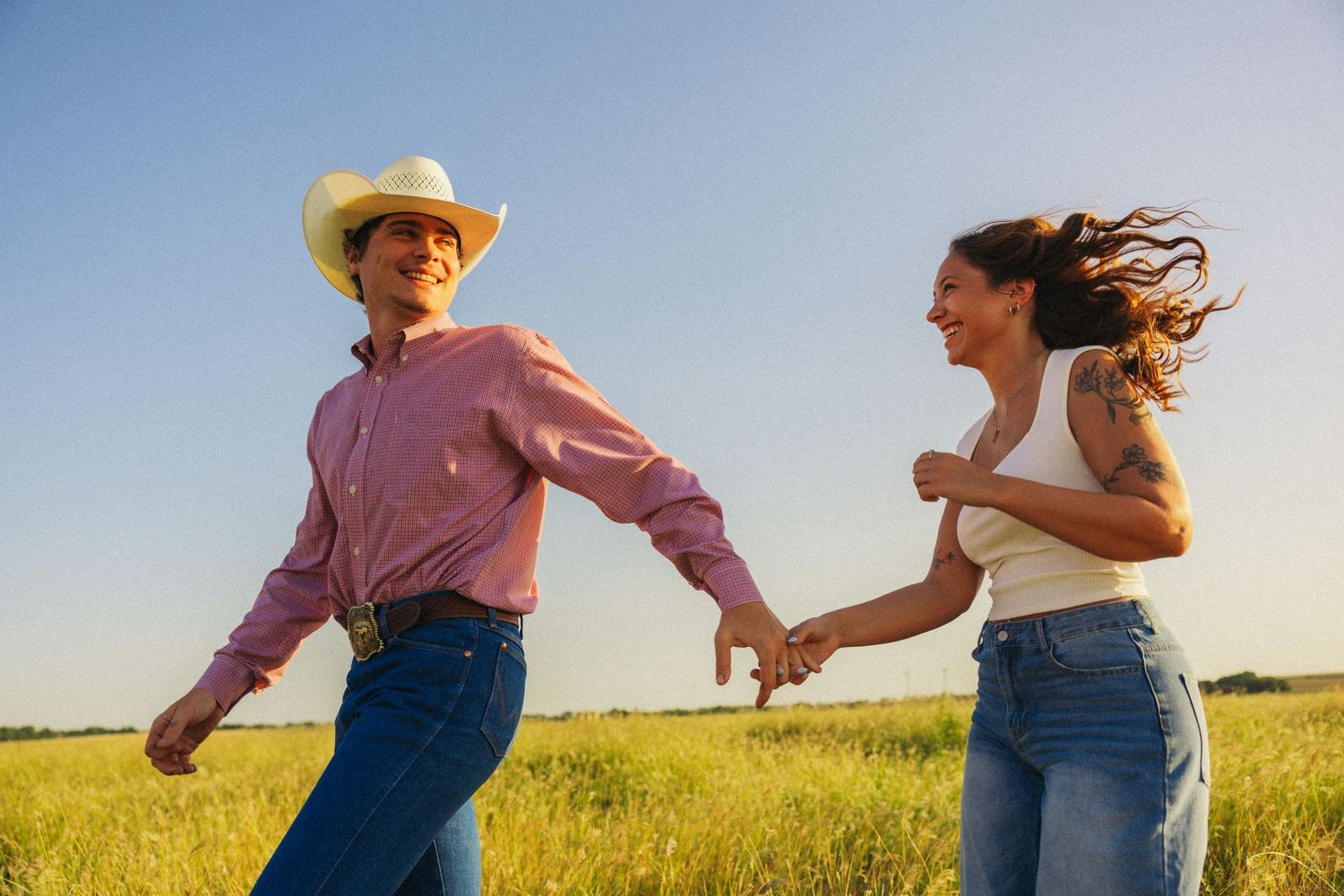 A smiling couple holds hands and walks through a sunny green field, the man in a cowboy hat and pink western shirt.