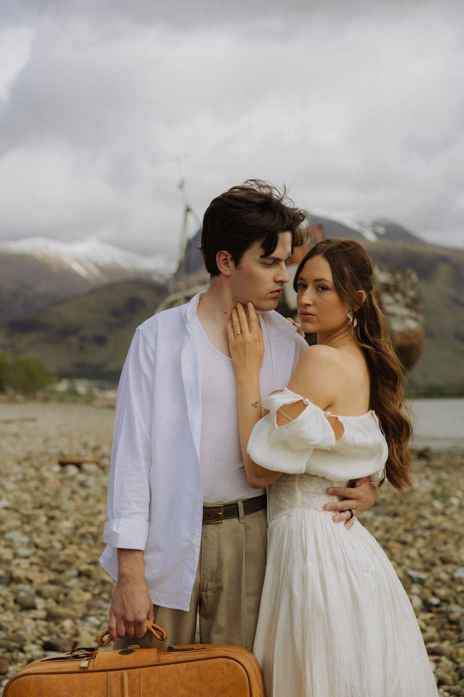 Romantic couple in white outfits embracing on rocky shore with mountains and vintage suitcase.