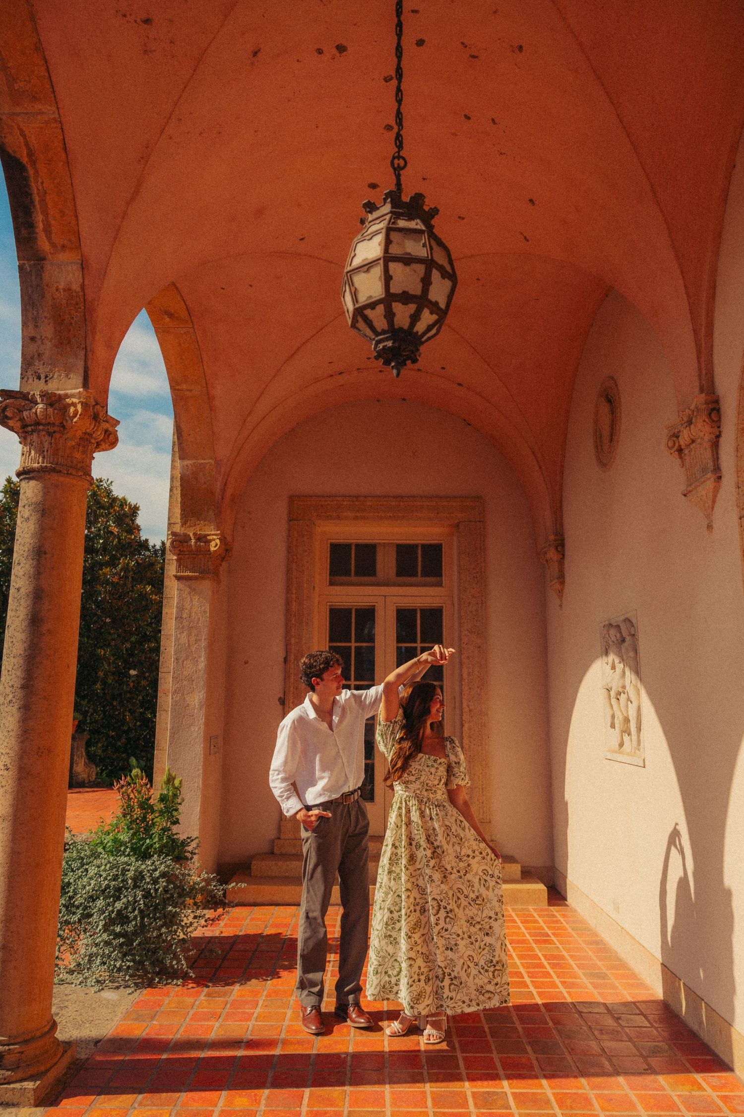Couple standing under arched colonnade with hanging lantern on terracotta tile floor at ornate Mediterranean estate.