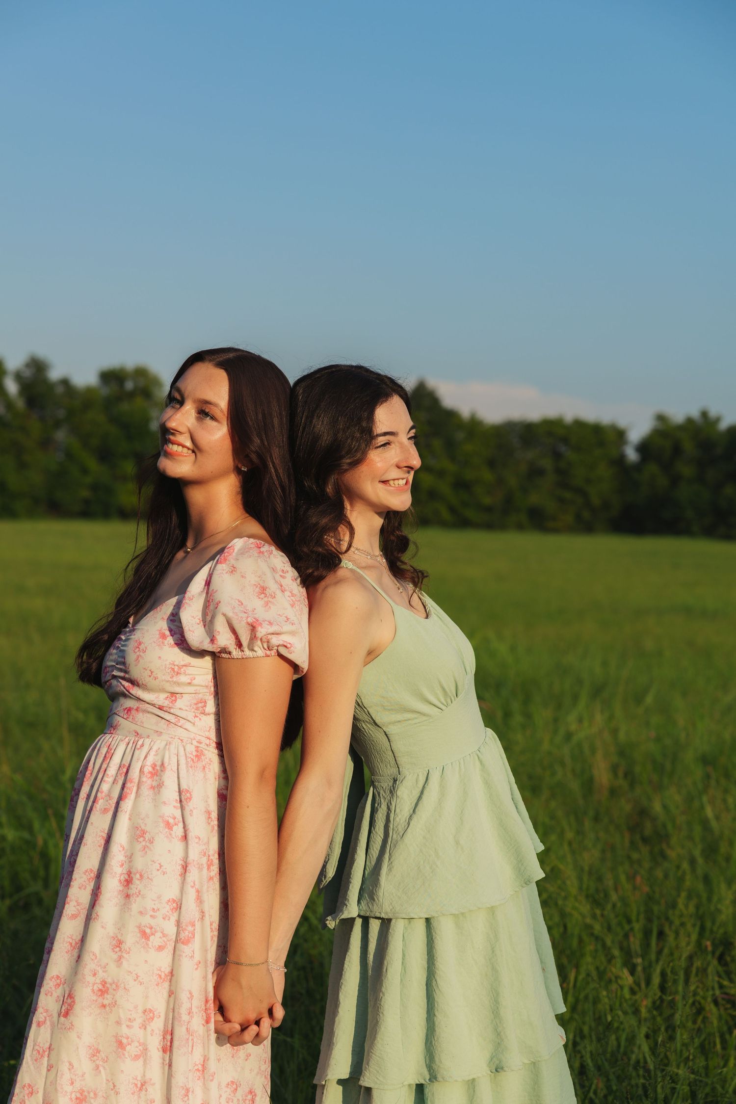 Two smiling young women in summer dresses holding hands back-to-back in a green field at golden hour.