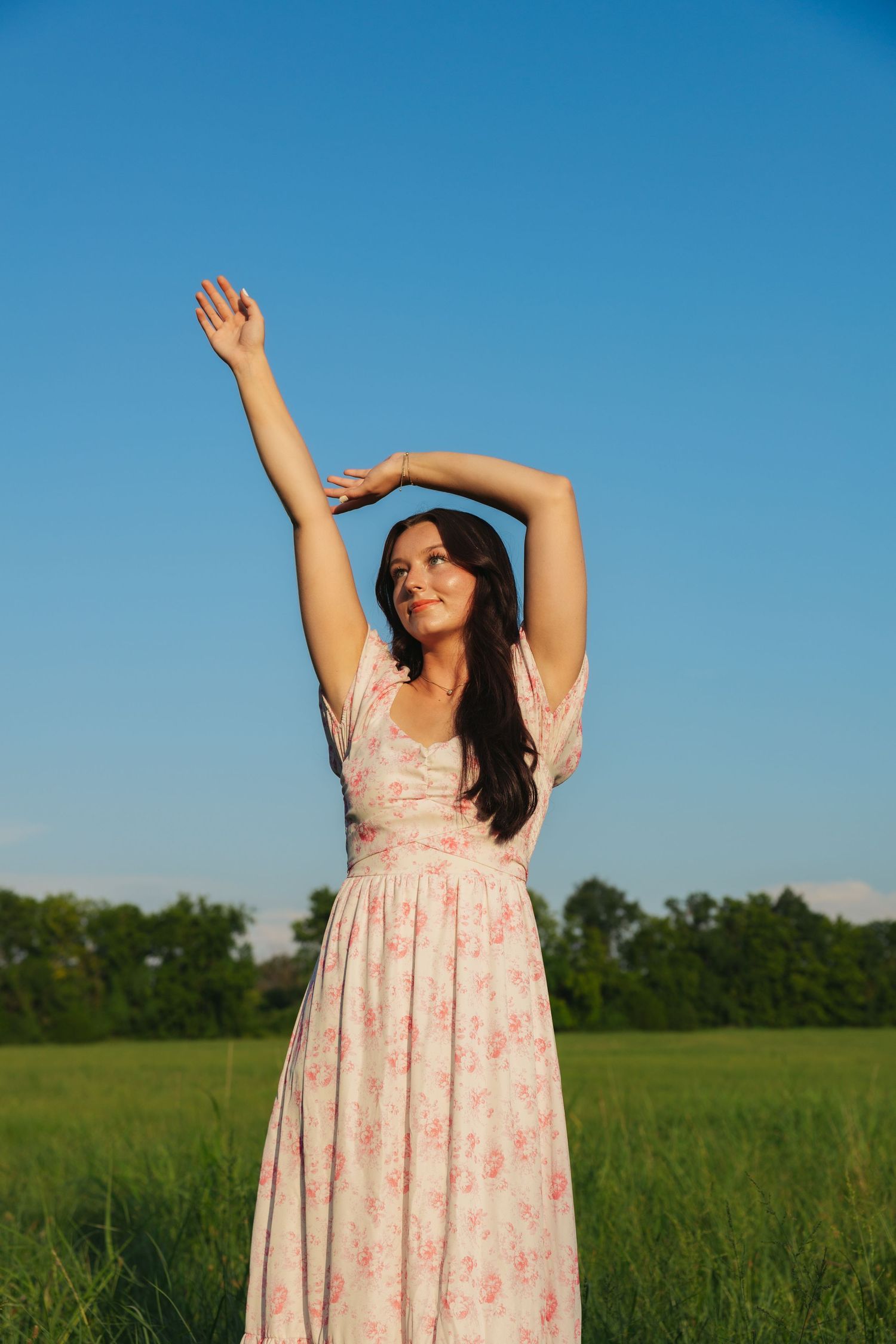 Young woman in floral pink dress with arms raised standing in green meadow under blue sky.