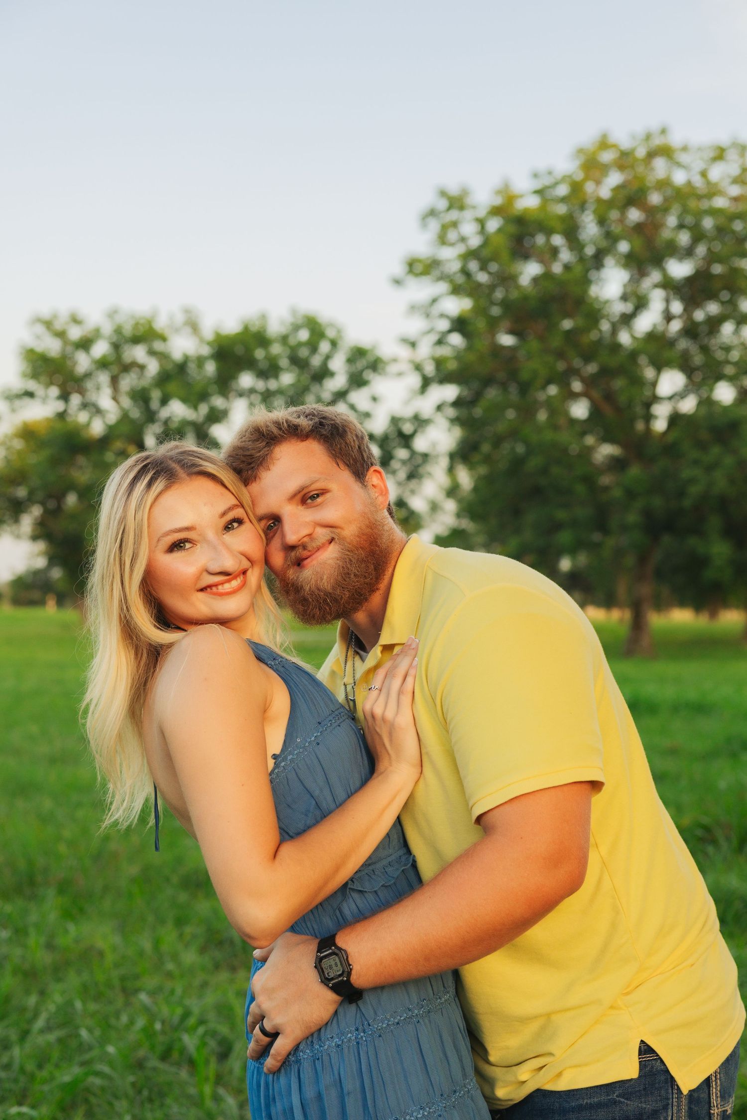 A bearded man in a yellow shirt tenderly embraces a blonde woman in a blue dress in a sunny green park setting.