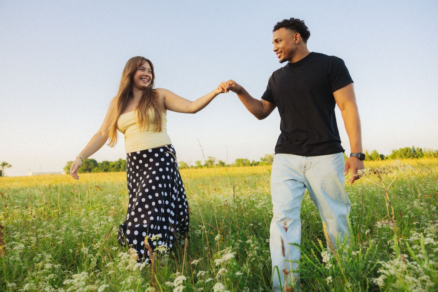 A joyful interracial couple holds hands and dances together in a blooming wildflower field under a clear blue sky.