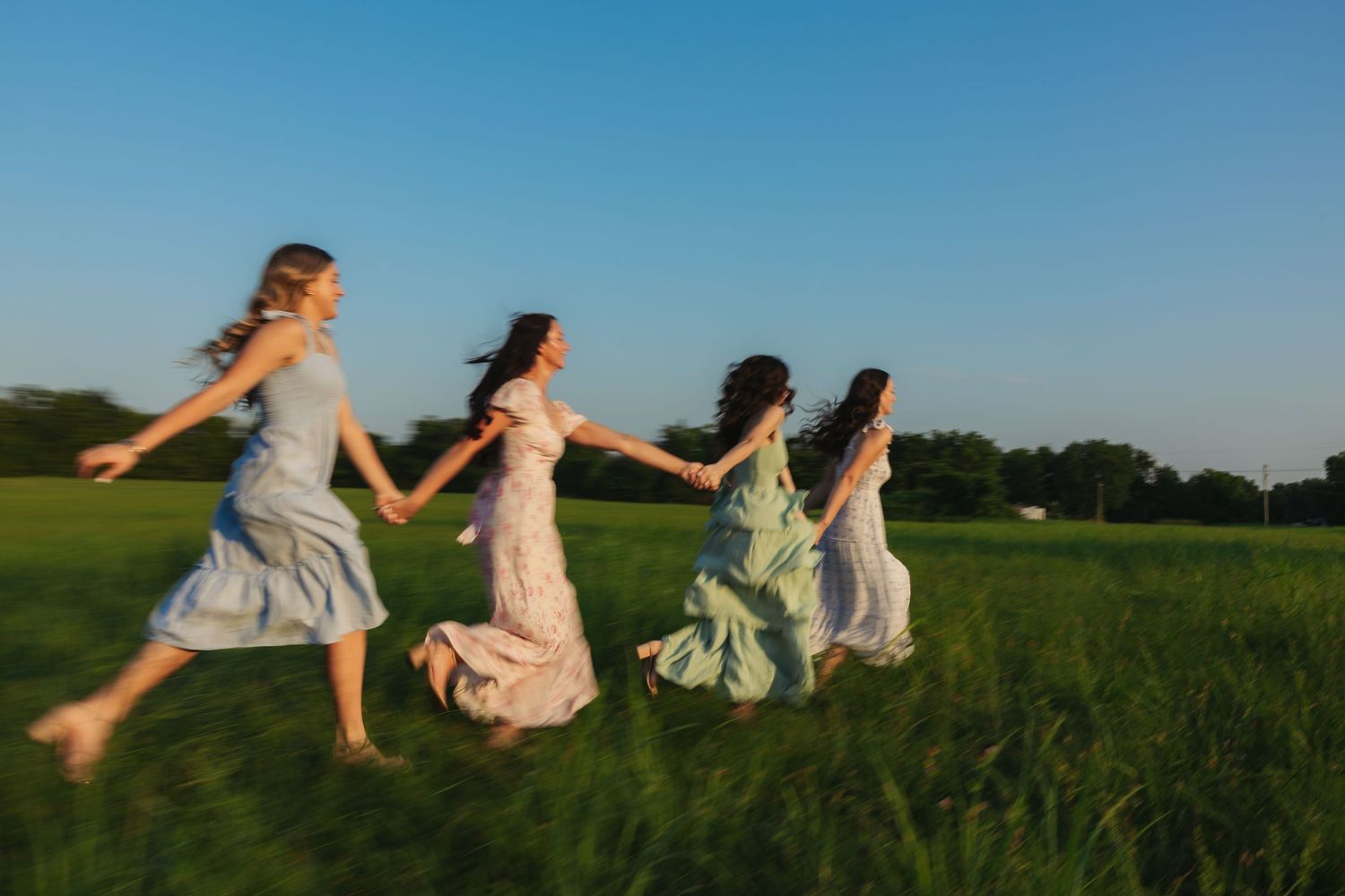 Four joyful women in colorful dresses running barefoot through a sunny meadow.