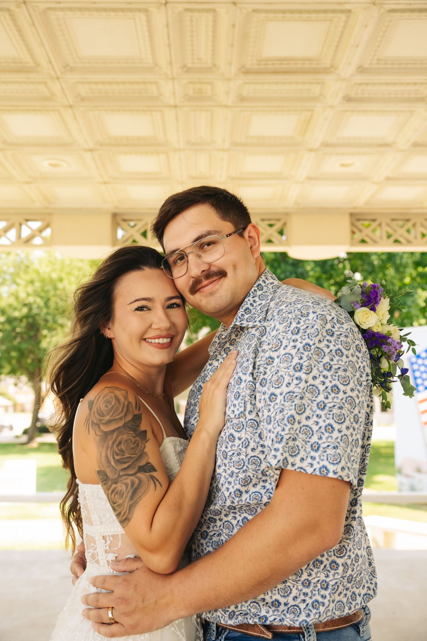 A smiling couple embraces outdoors, the woman wearing a white dress with rose tattoos, the man in a floral shirt with glasses.
